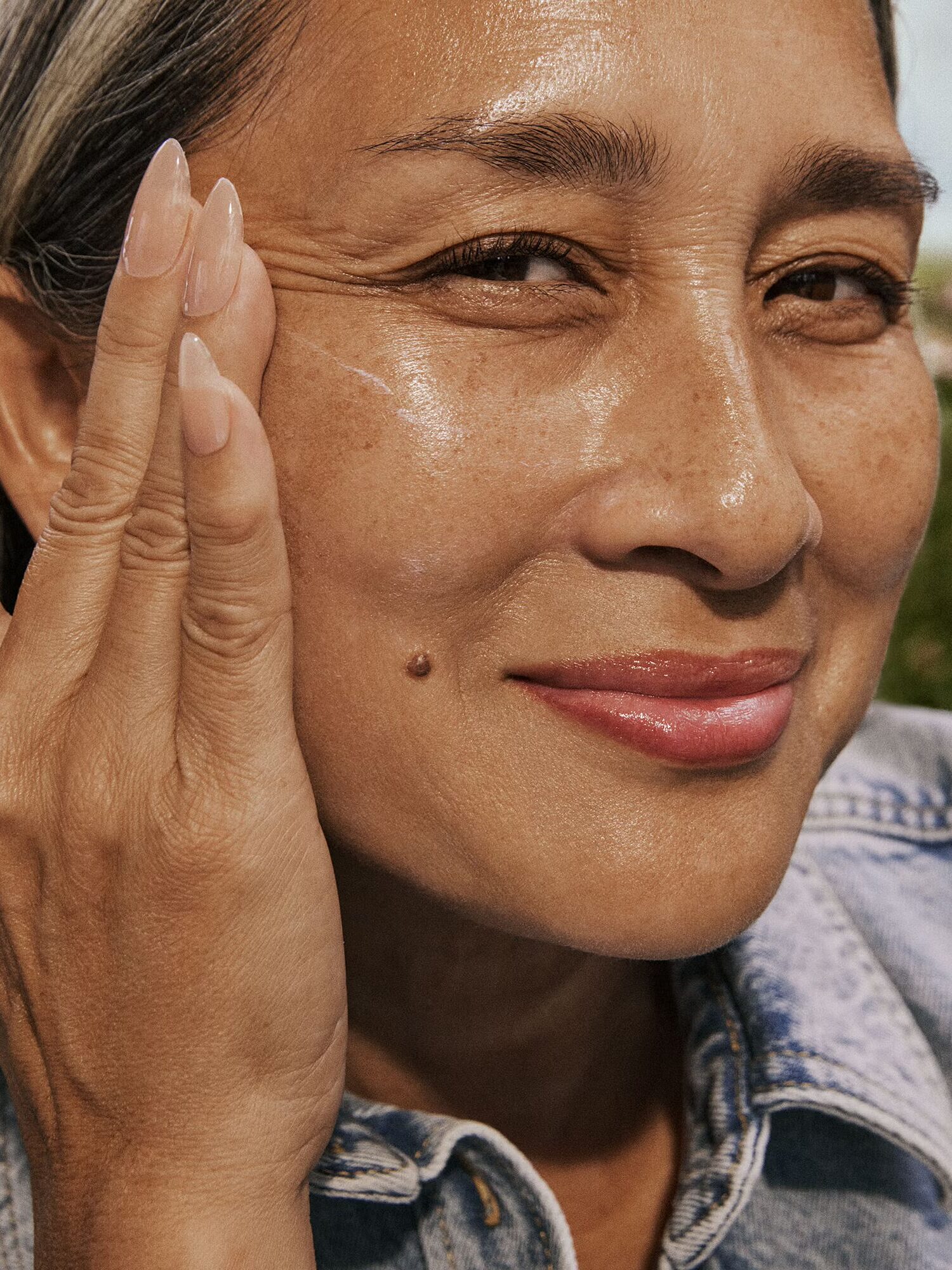 Woman with long fingernails smiling, touching her face with one hand, wearing a denim jacket, outdoors.