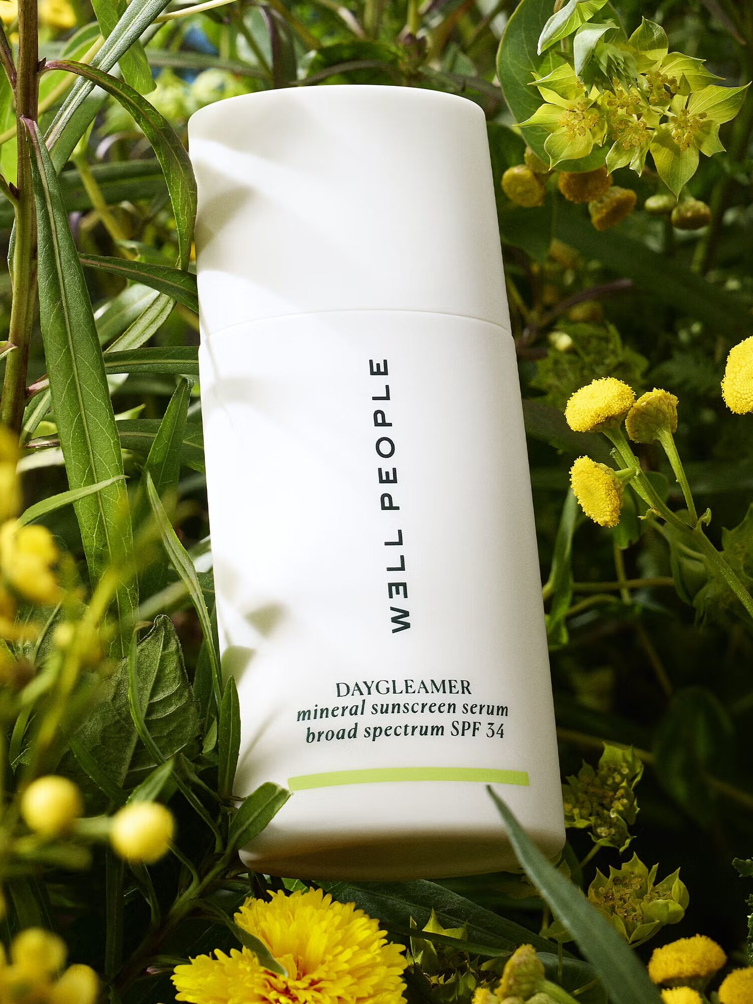 A bottle of Well People Dayglow Mineral Sunscreen SPF 35 stands among yellow and green flowers and leaves.
