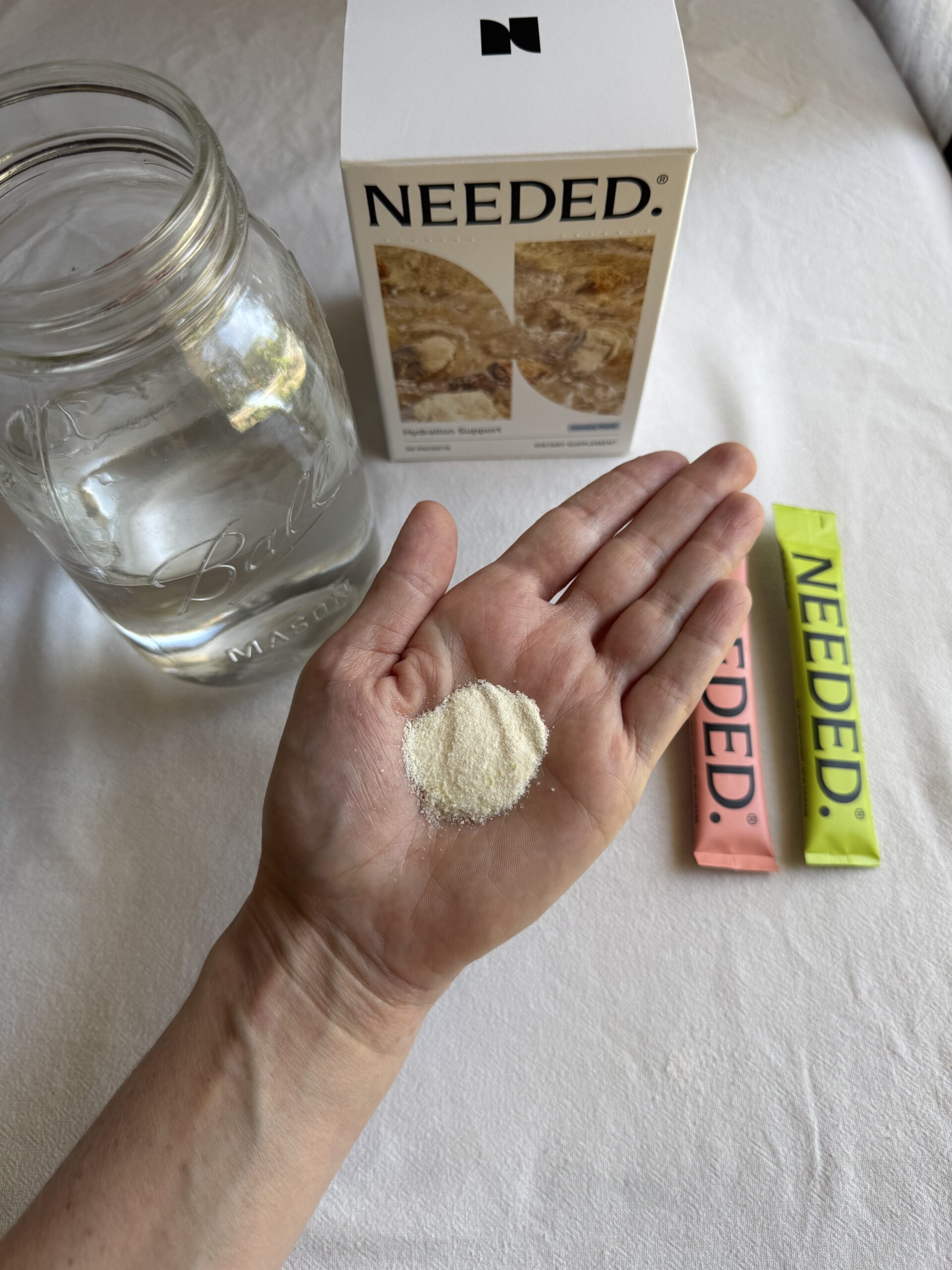 A hand holds a small mound of white powder in front of a mason jar of water, a box labeled "Needed," and two colored supplement packets.
