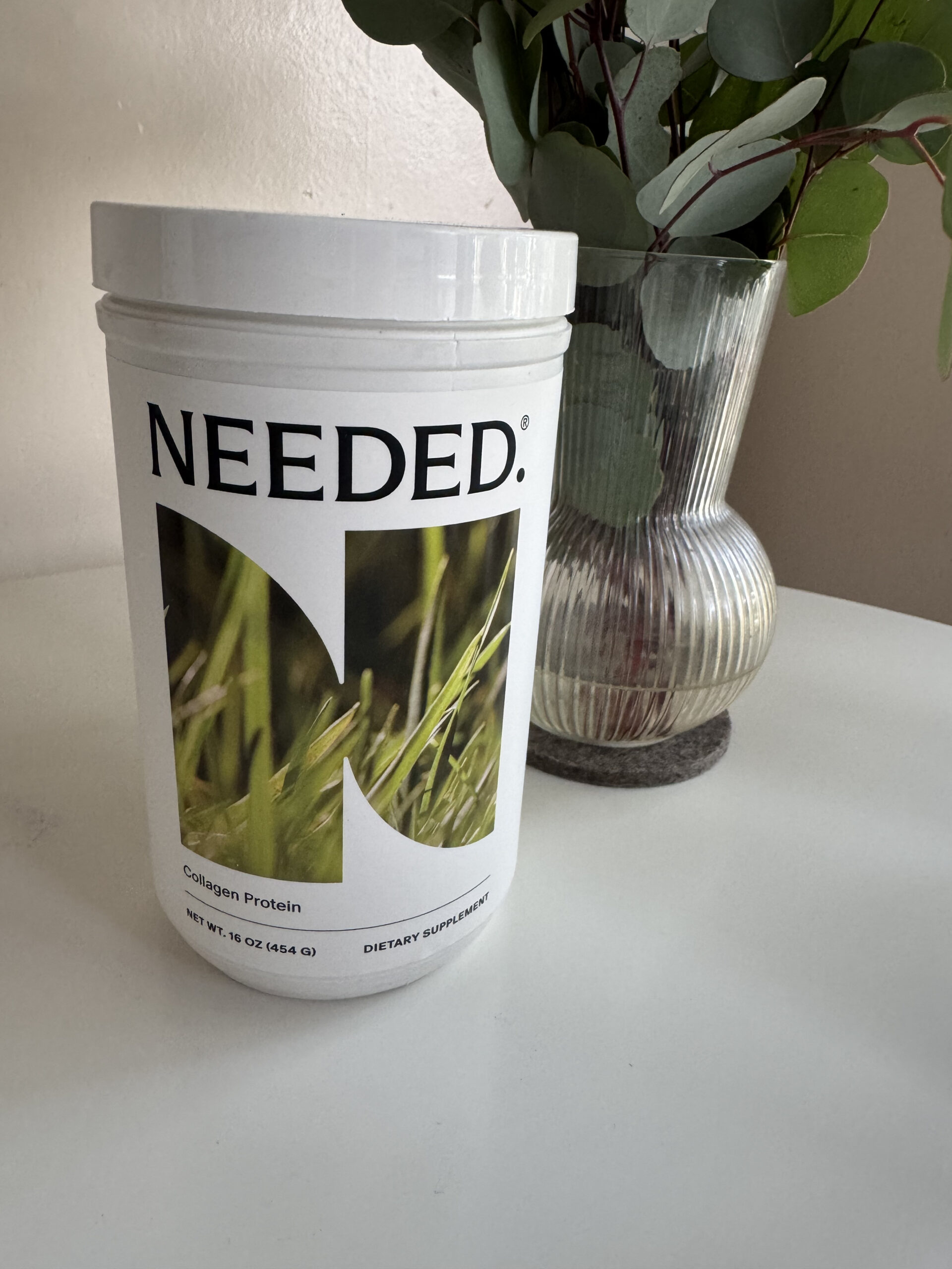 A white container labeled "NEEDED." with an image of grass and text "Collagen Protein," sits on a white surface next to a ribbed glass vase with greenery.