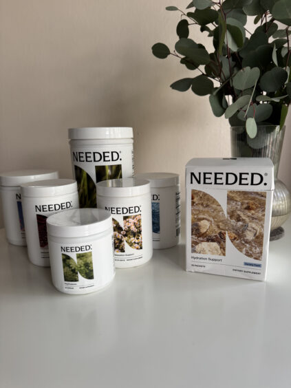 Seven supplement containers labeled "NEEDED." are displayed on a surface with a leafy plant in a vase behind them.