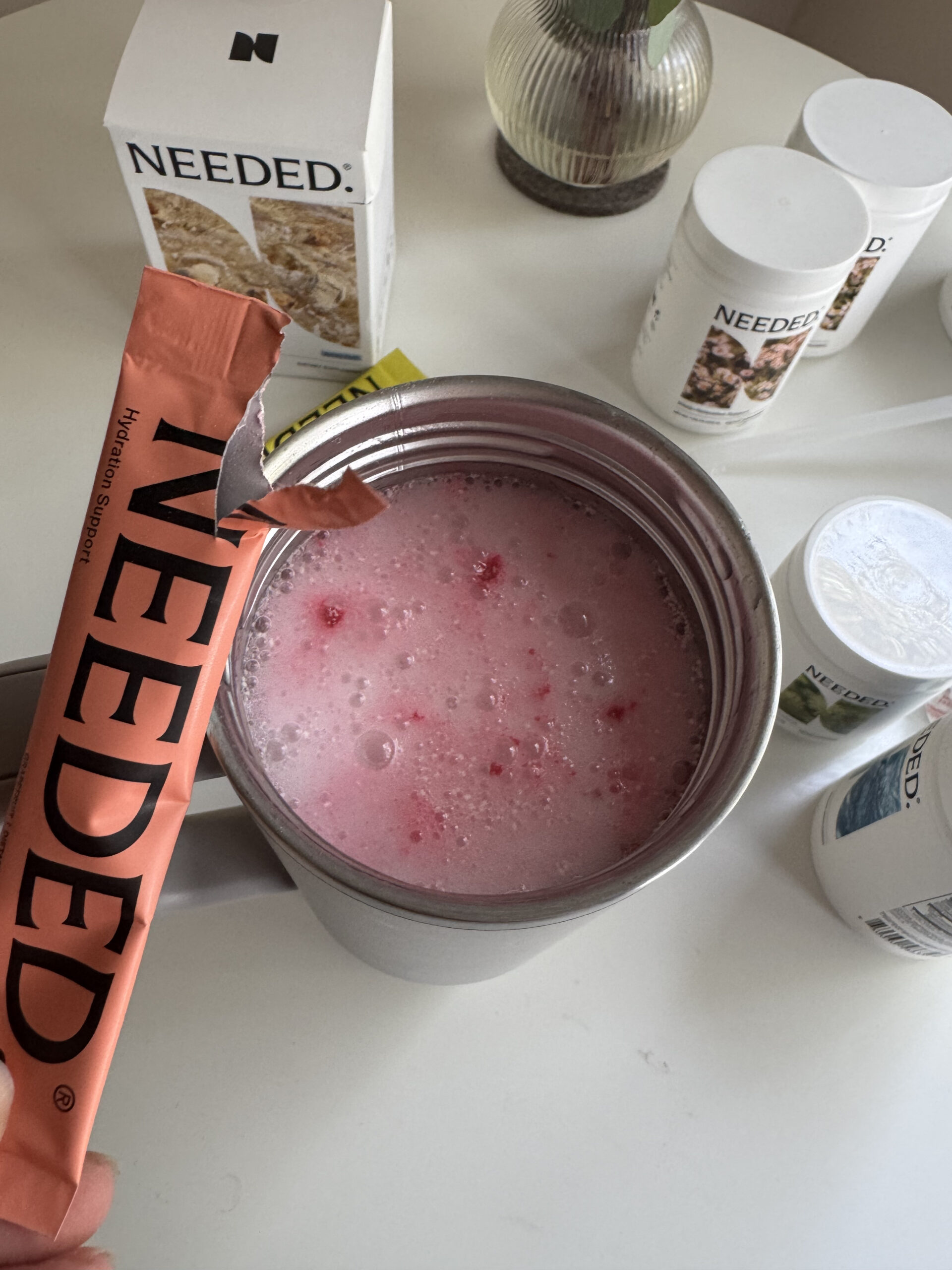 A frothy pink drink in a cup, surrounded by containers labeled "Needed." A hand holds an opened "Needed" packet above the cup.