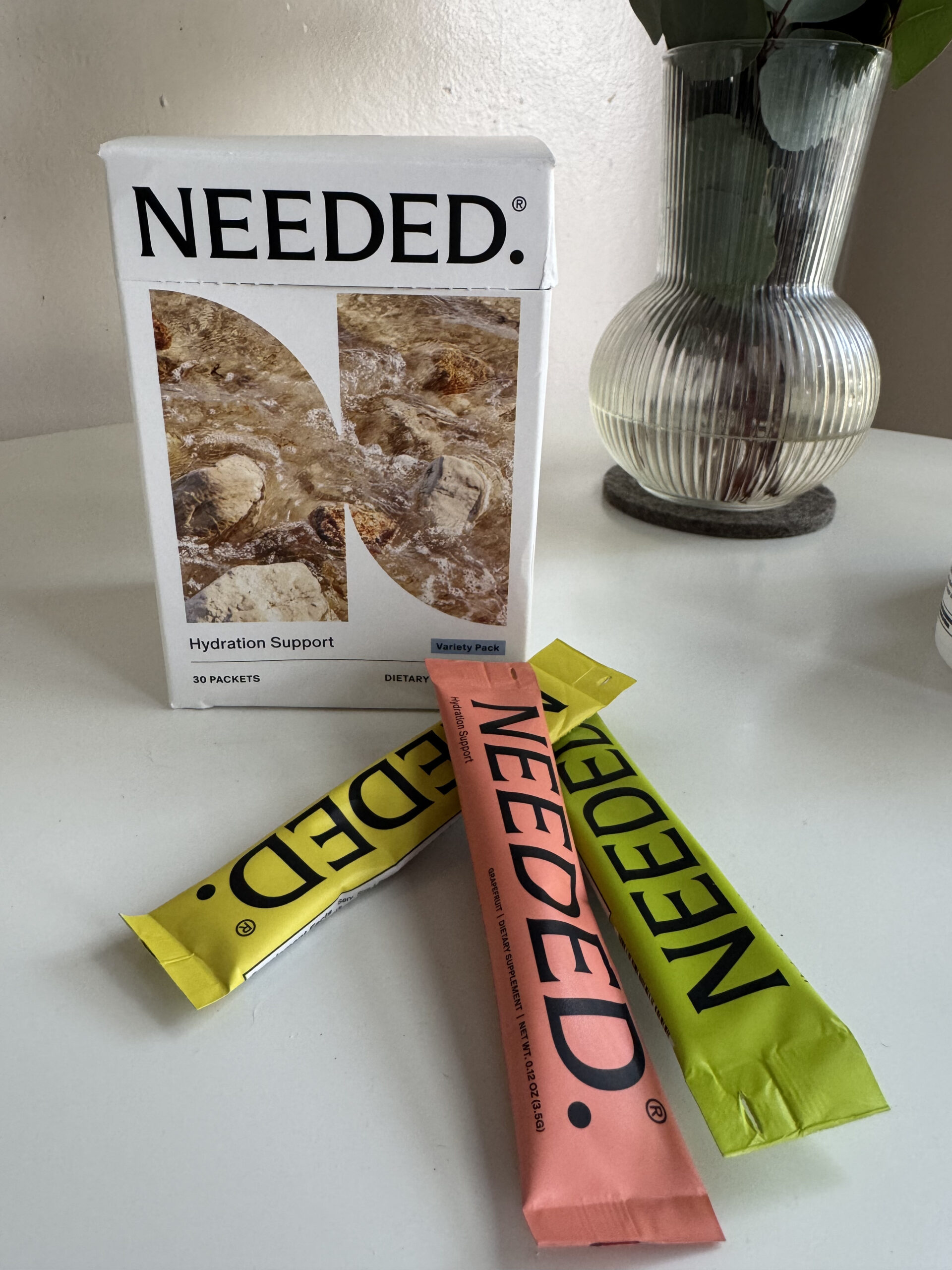 Box of NEDED Hydration Support on a table with three colorful sachets in front, next to a vase with green leaves.