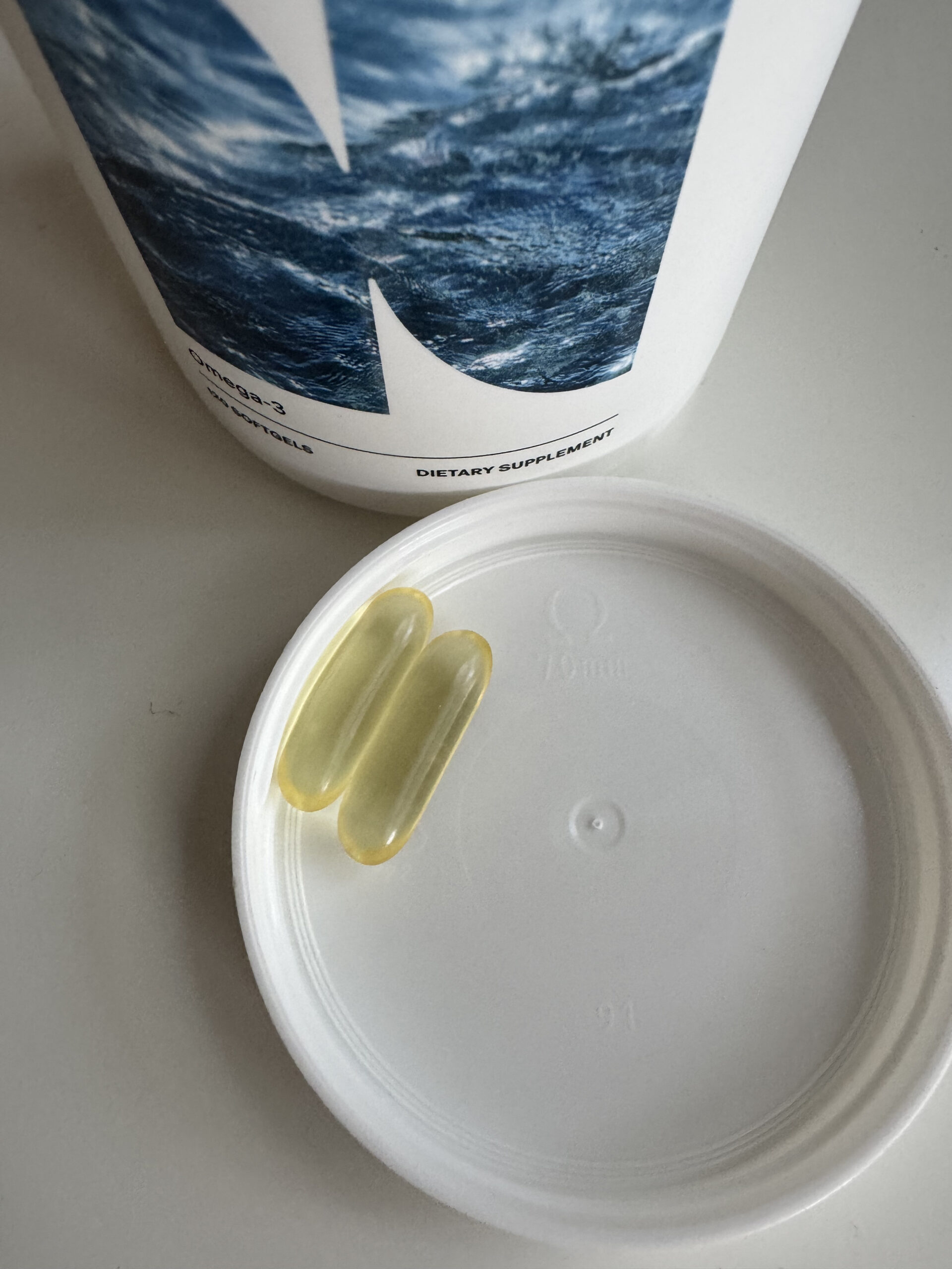 Two yellow capsules on a white plastic lid, next to a container with a label featuring a water image and text "Dietary Supplement.