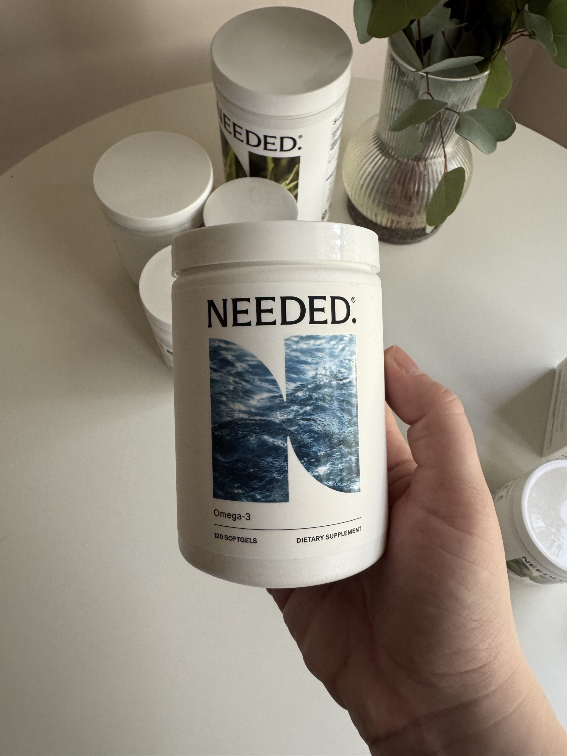 A hand holds a container of Needed Omega-3 dietary supplement softgels. Other similar containers are blurred in the background on a round table.