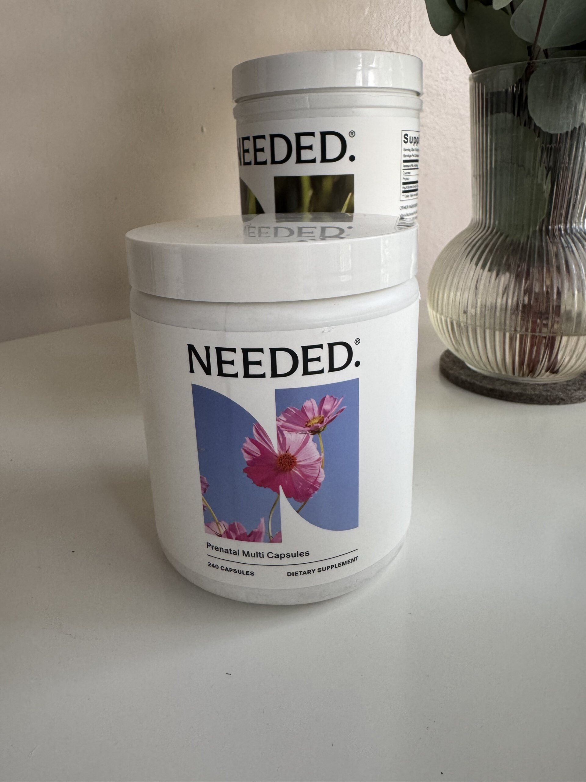 A container of "Needed" prenatal multi capsules is on a table next to a vase and another similar container.