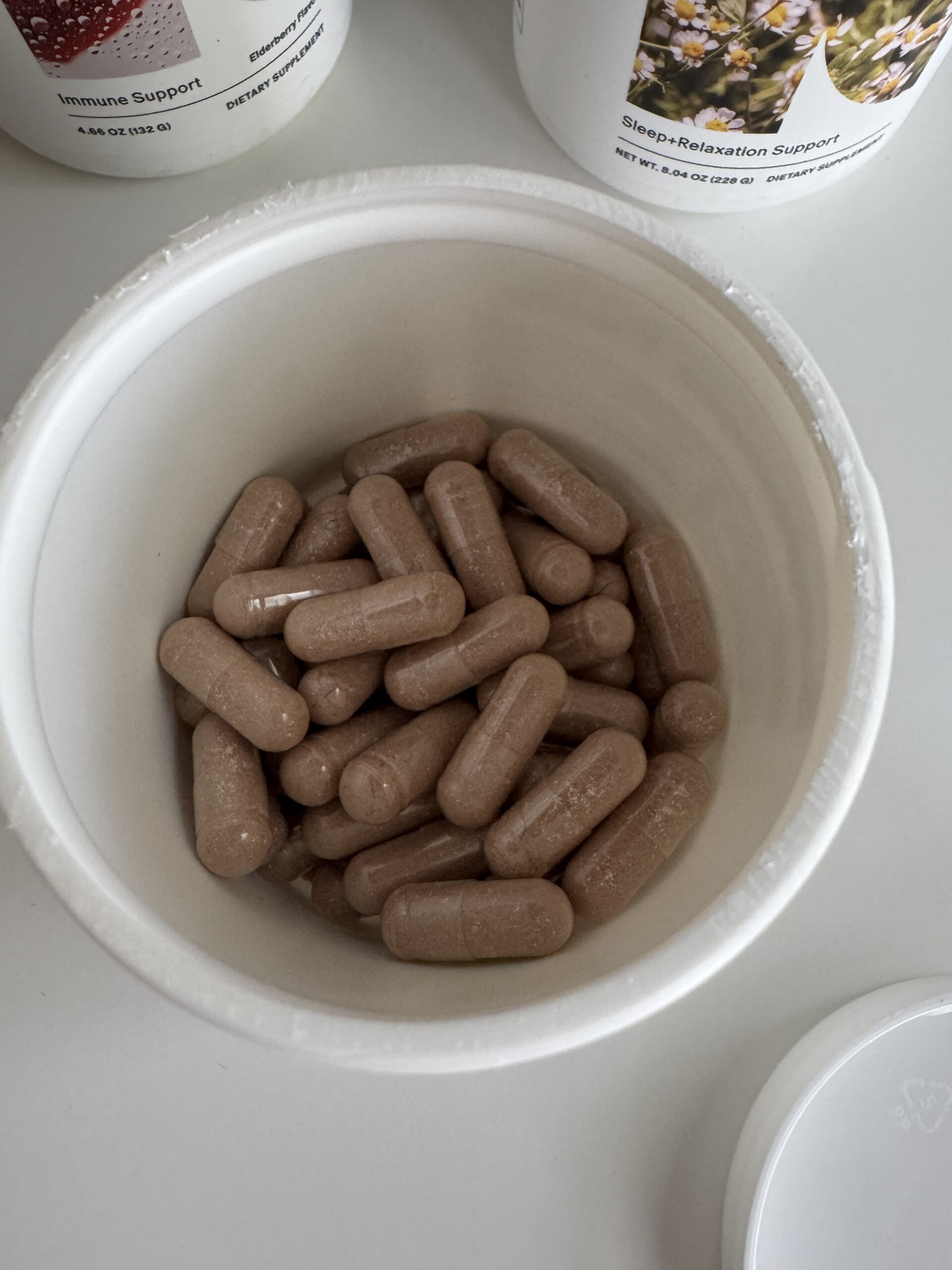 Open container filled with brown capsules on a white surface, with closed containers labeled "Immune Support" and "Sleep-Relaxation Support" in the background.