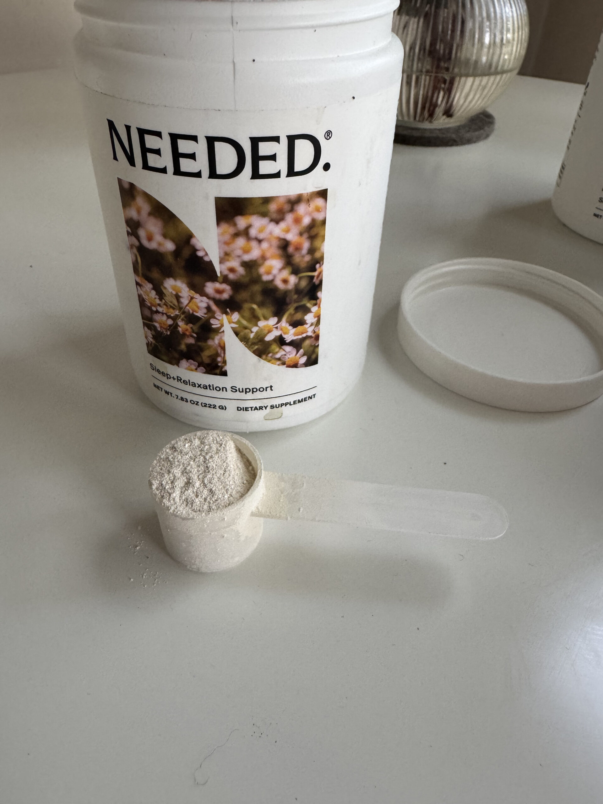 A white container labeled "NEEDED" with a floral design is open on a table. A scoop of white powder is beside it, with some powder spilled on the surface.