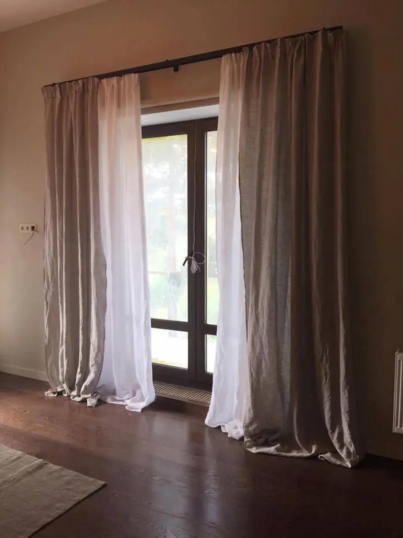 Large window with sheer white curtains on a dark rod, overlooking a greenery outside. Brown hardwood floor and beige walls surround the area.