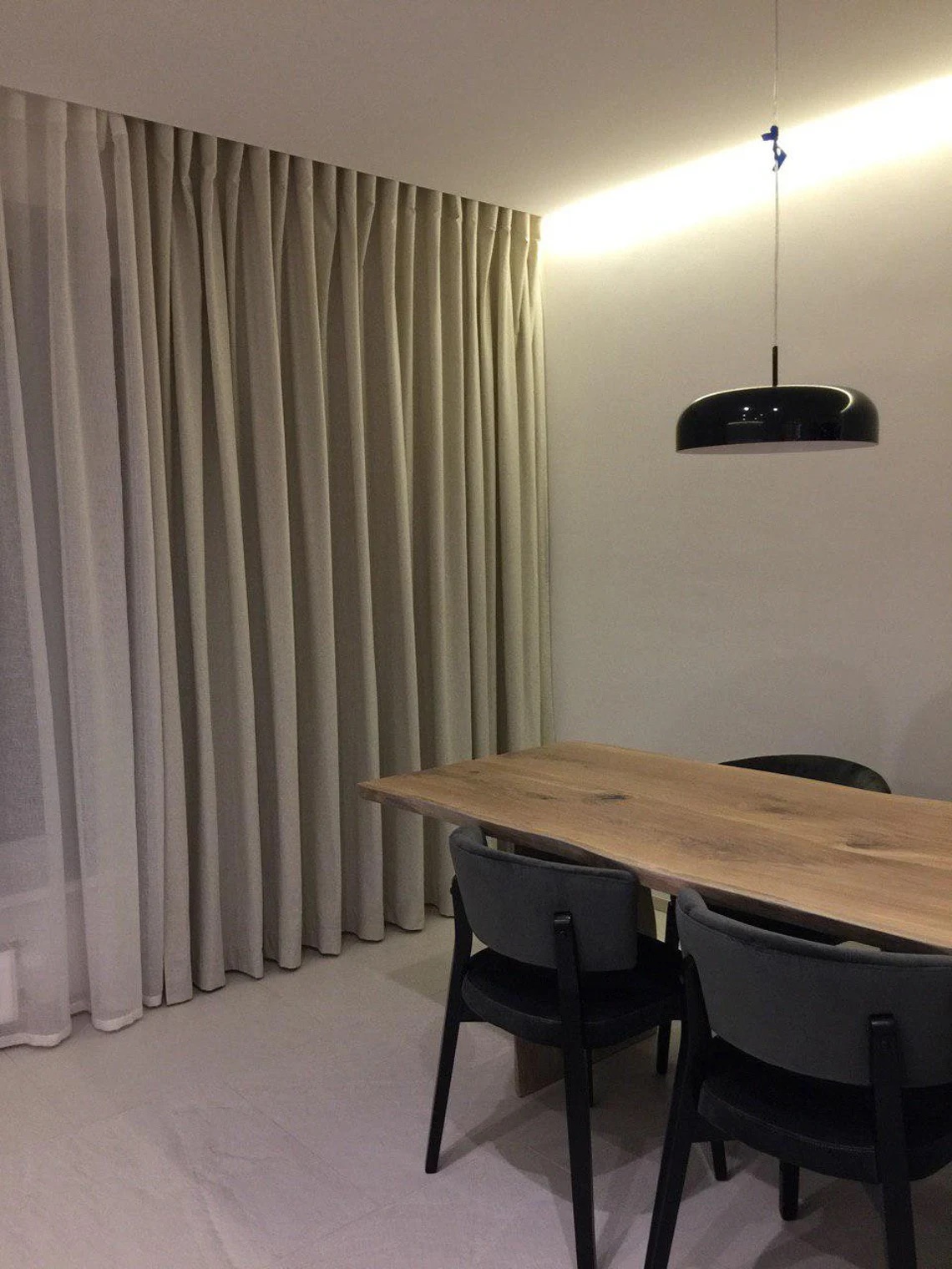 A dining area with a wooden table, four dark chairs, sheer curtains, and a black pendant light hanging from the ceiling.