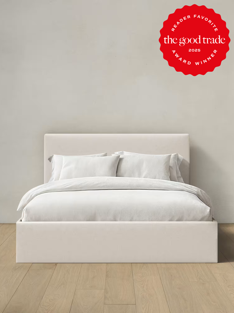 A minimalist white bed with white bedding stands against a plain light-colored wall on a light wood floor. A red "Reader Favorite" award badge from The Good Trade 2025 is in the top right corner.