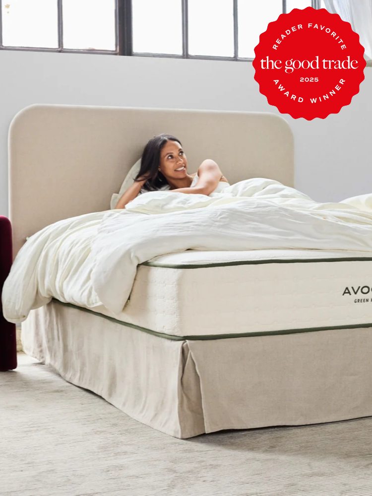 A woman sits up in bed, smiling, covered by white bedding on an Avocado Green mattress in a bright, modern bedroom. A red award badge is displayed in the top right corner.