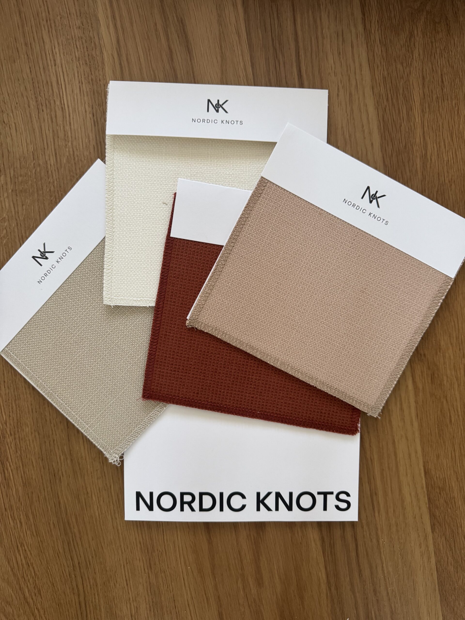 Four fabric swatches with labels reading "Nordic Knots" are arranged on a wood surface. The swatches are in white, beige, light brown, and rust shades.