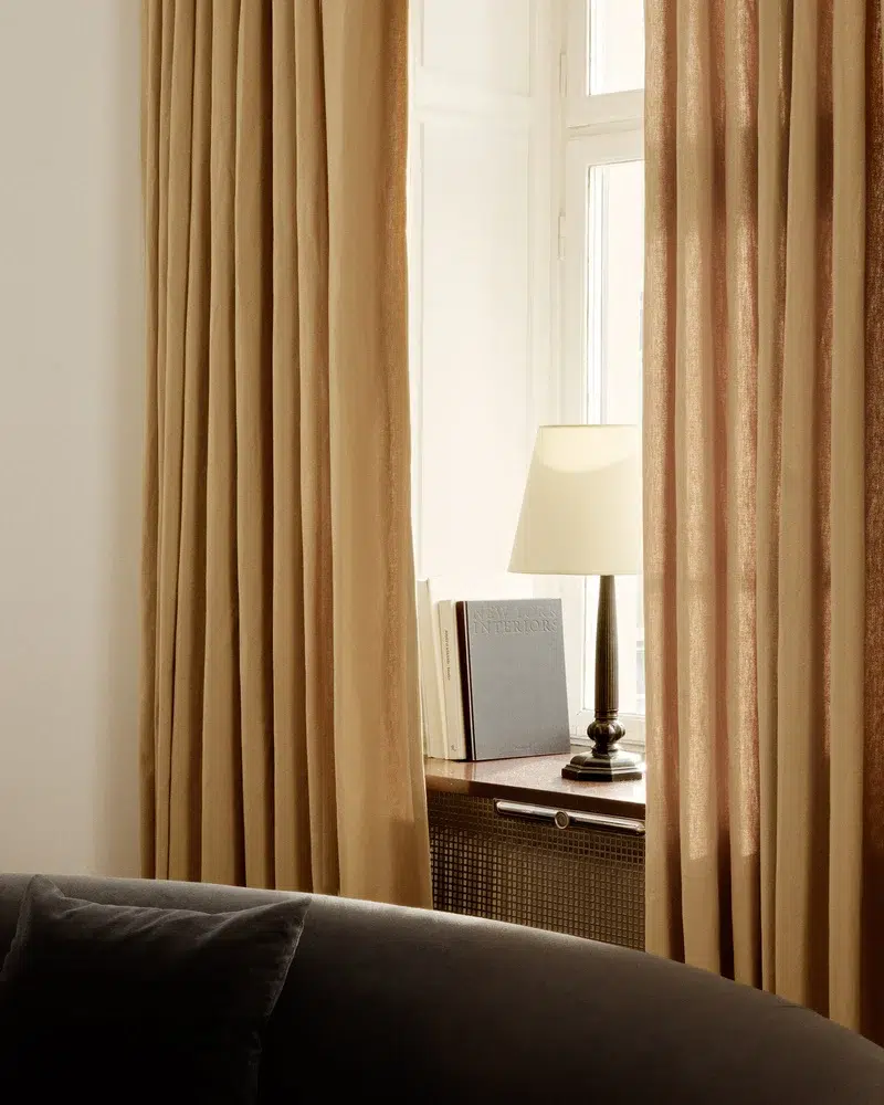 A lamp and books on a table by a window with closed beige curtains. A portion of a dark cushioned chair is visible.