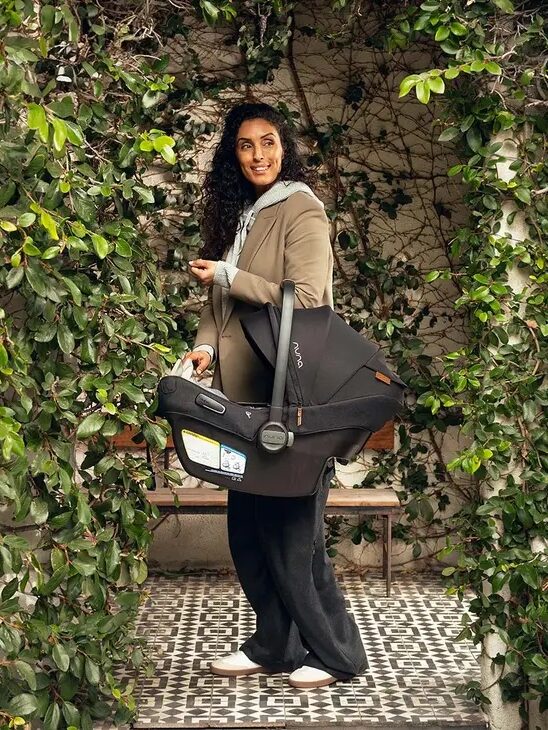 A person stands outdoors holding a baby car seat. They are surrounded by green leafy plants and patterned tiles underfoot.