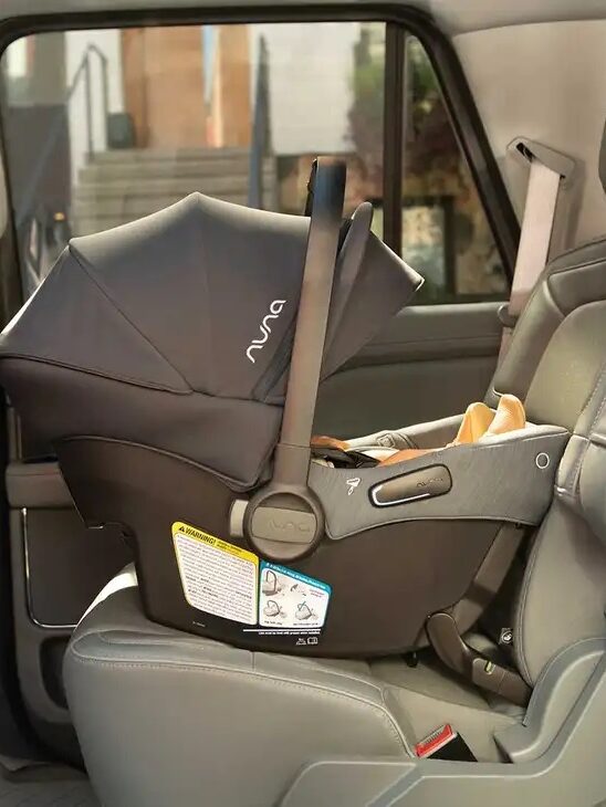A black baby car seat is securely installed in the backseat of a vehicle, with a sunshade extended.
