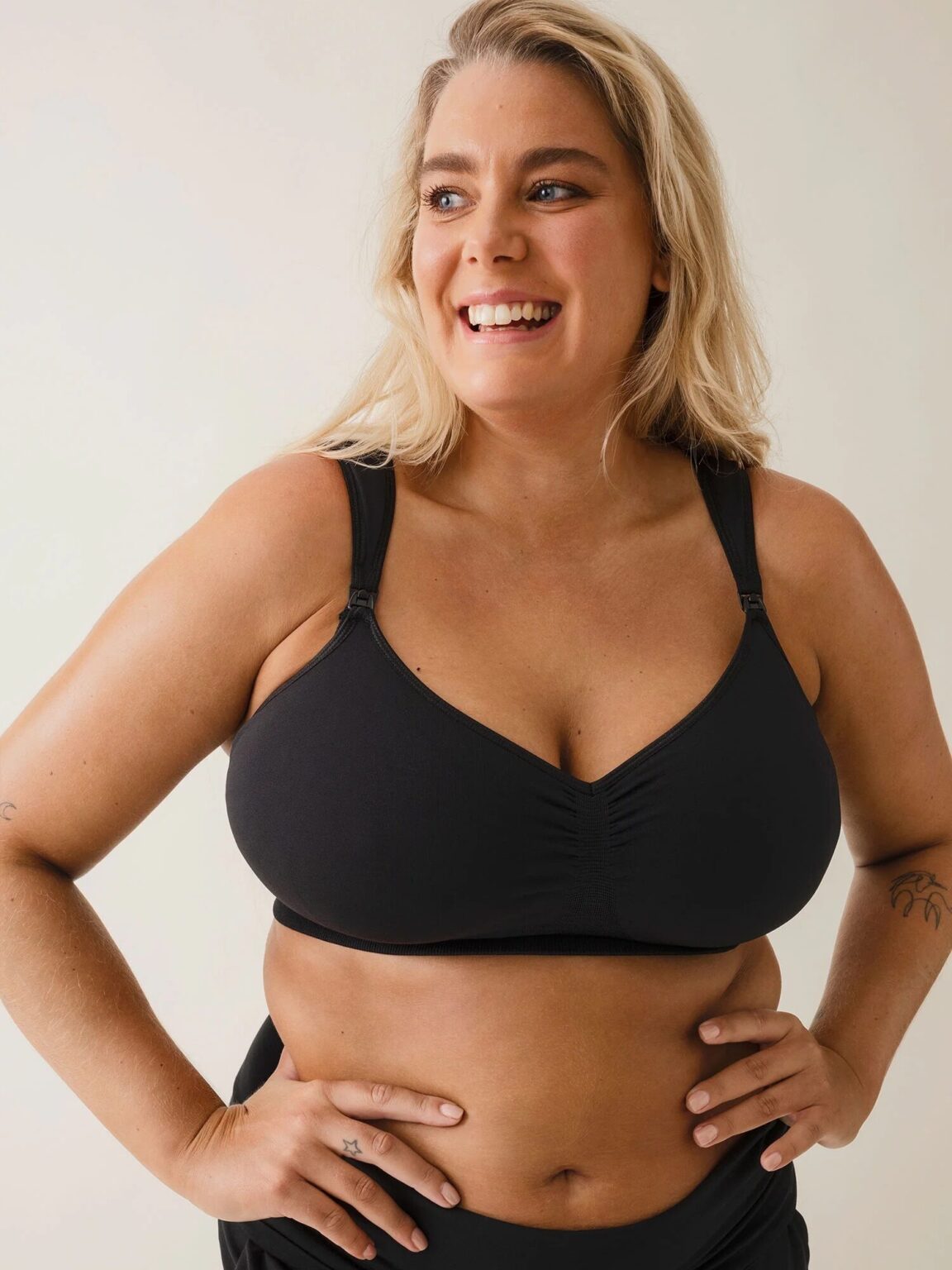 A smiling woman with long blonde hair wears a black sports bra, standing with hands on hips against a neutral background.