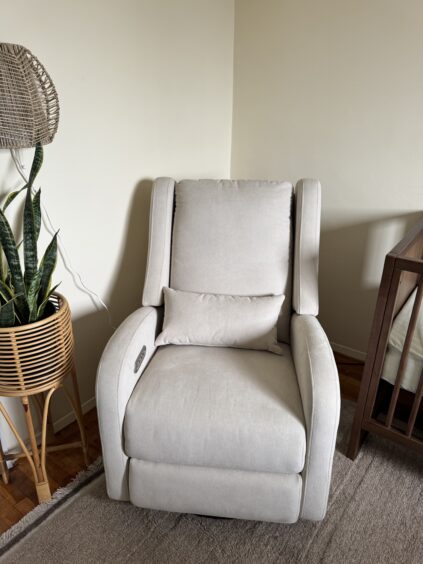 A light gray upholstered armchair with a matching pillow sits on a rug next to a wooden plant stand and a crib in a corner of a room.