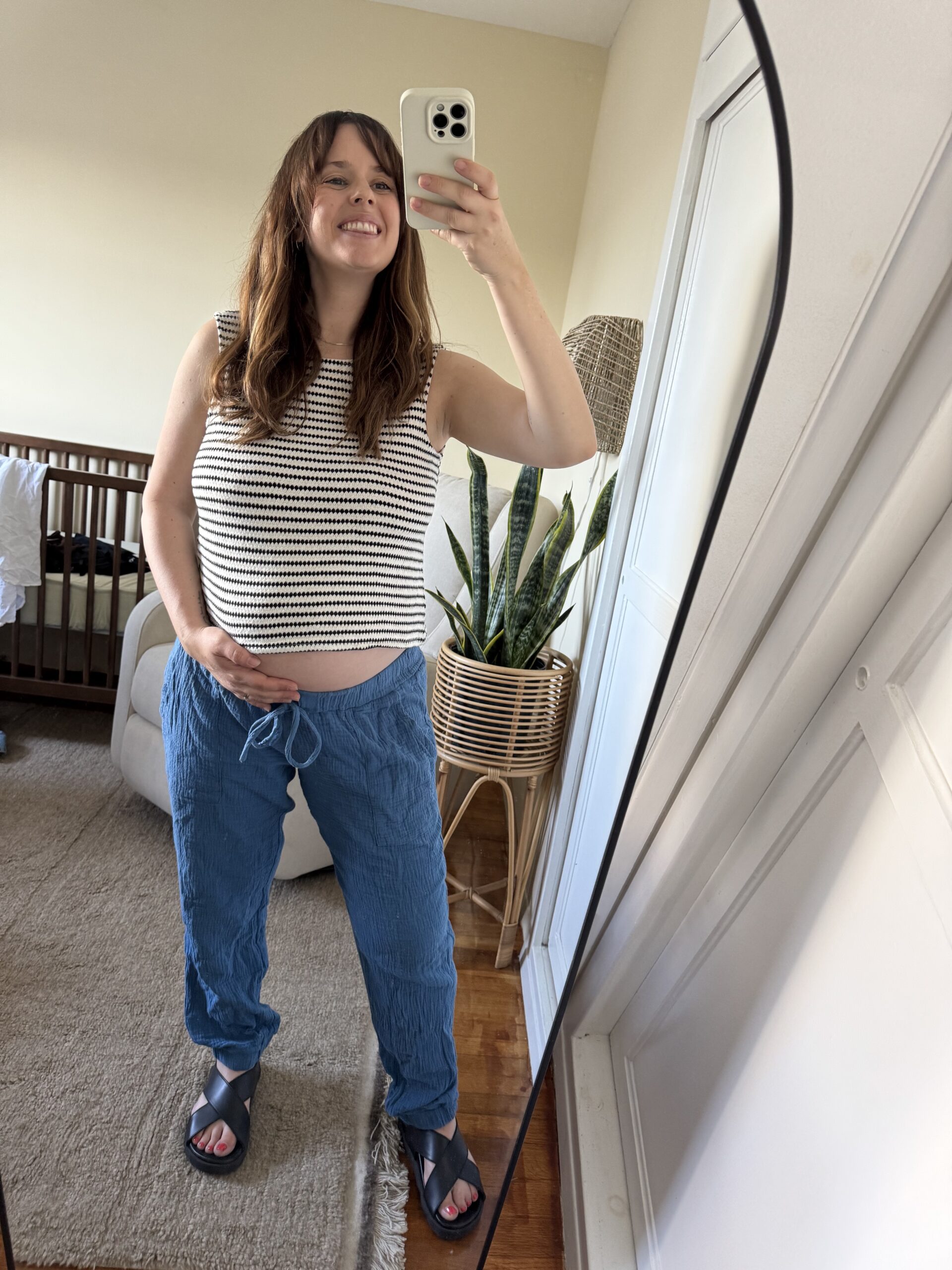 Woman taking a mirror selfie in a bedroom, smiling, with one hand on her pregnant belly. She is wearing a striped sleeveless shirt, blue pants, and sandals.