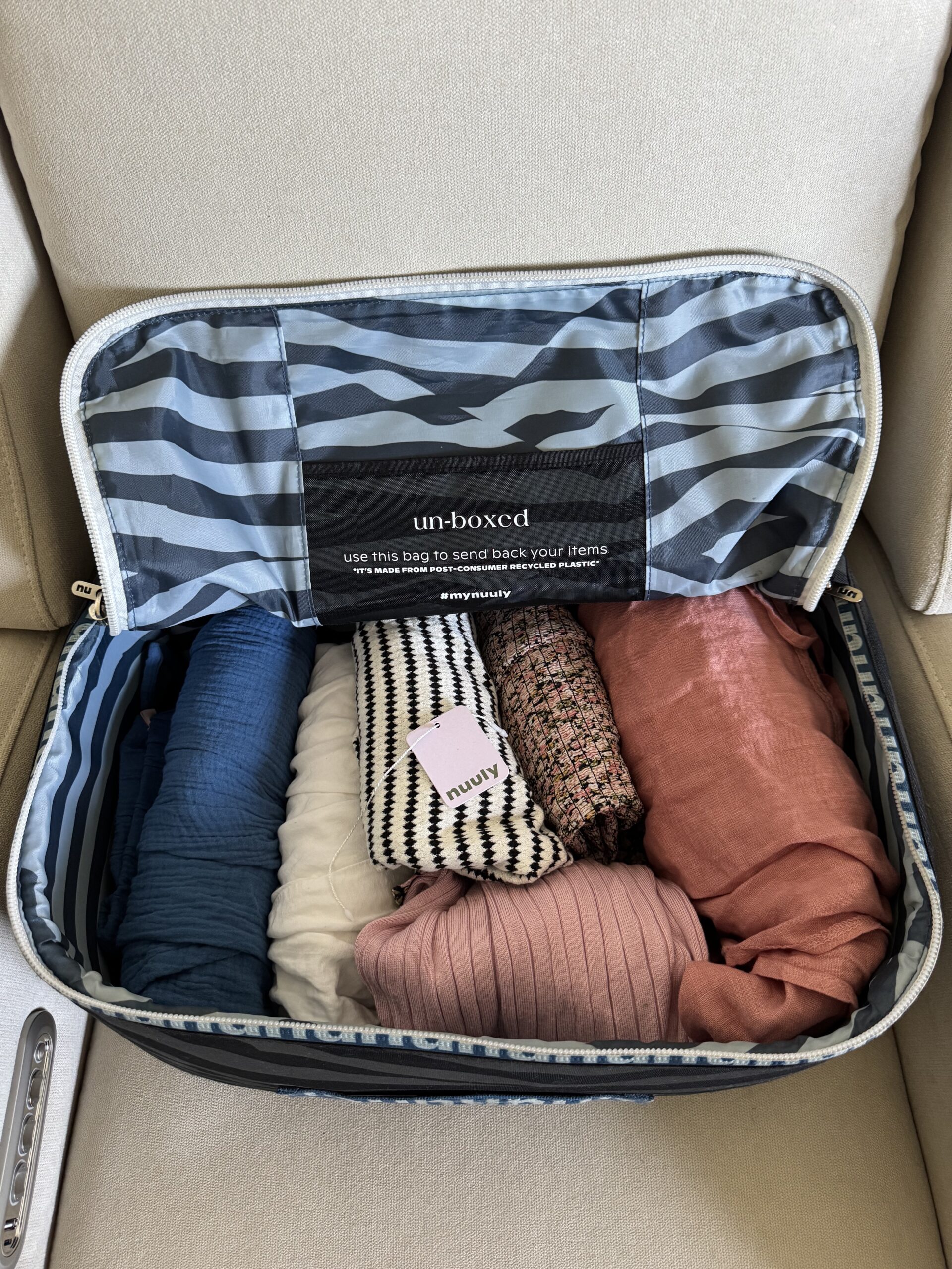 An open suitcase on a beige chair holds neatly rolled clothing in blue, white, black-and-white, pink, and patterned fabrics, with a small card placed on top.