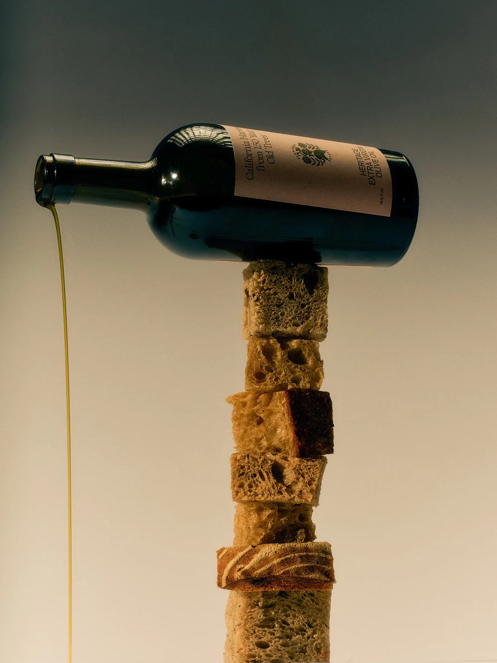 A bottle of olive oil balanced on a vertical stack of bread pieces, with oil pouring out.