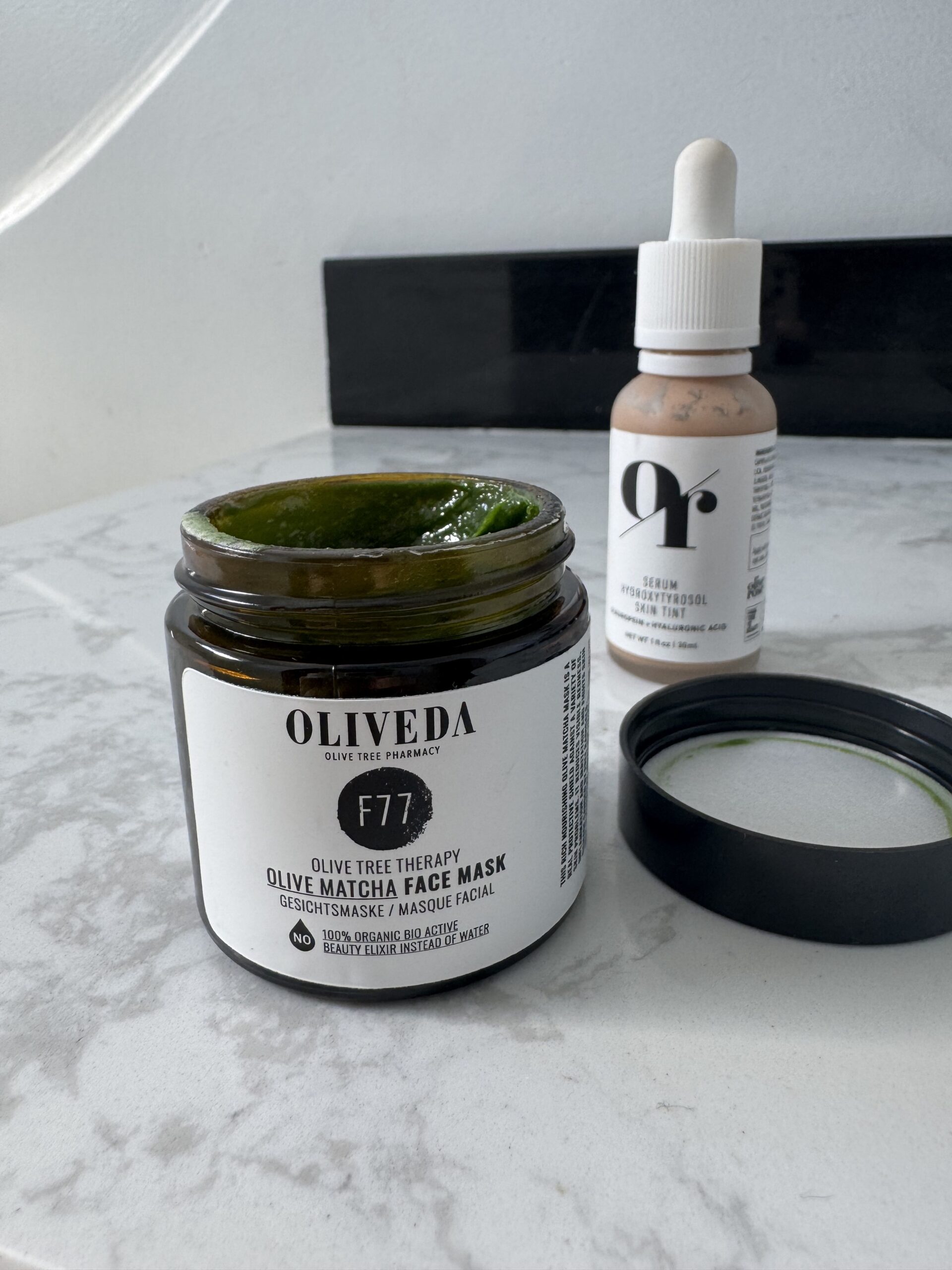 A jar of Oliveda face mask and a dropper bottle of serum sit on a marble surface.