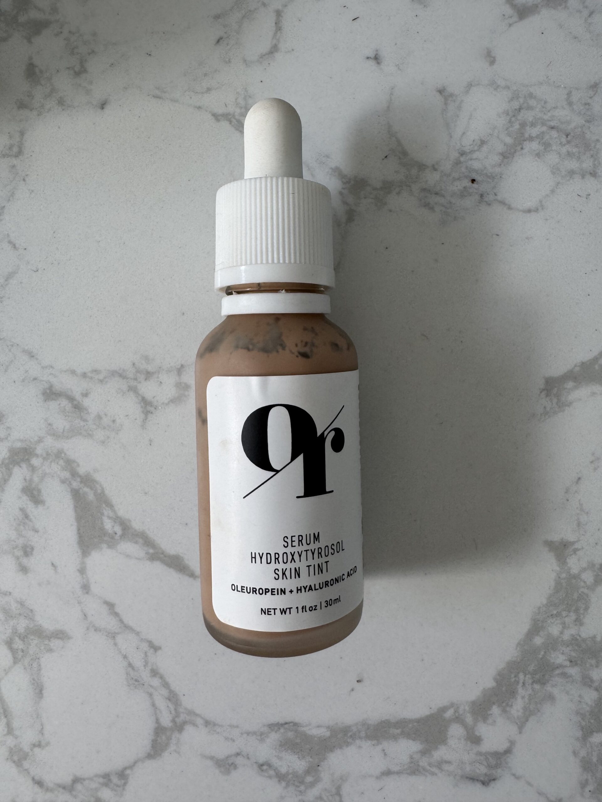 A small bottle with a dropper cap labeled "Serum Hydroxytyrosol Skin Tint" on a marble surface.