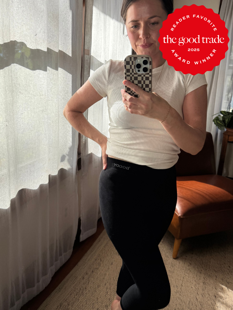 A person stands indoors taking a mirror selfie, wearing a white top and black leggings. A red "The Good Trade 2025 Award Winner" badge appears in the top right corner.