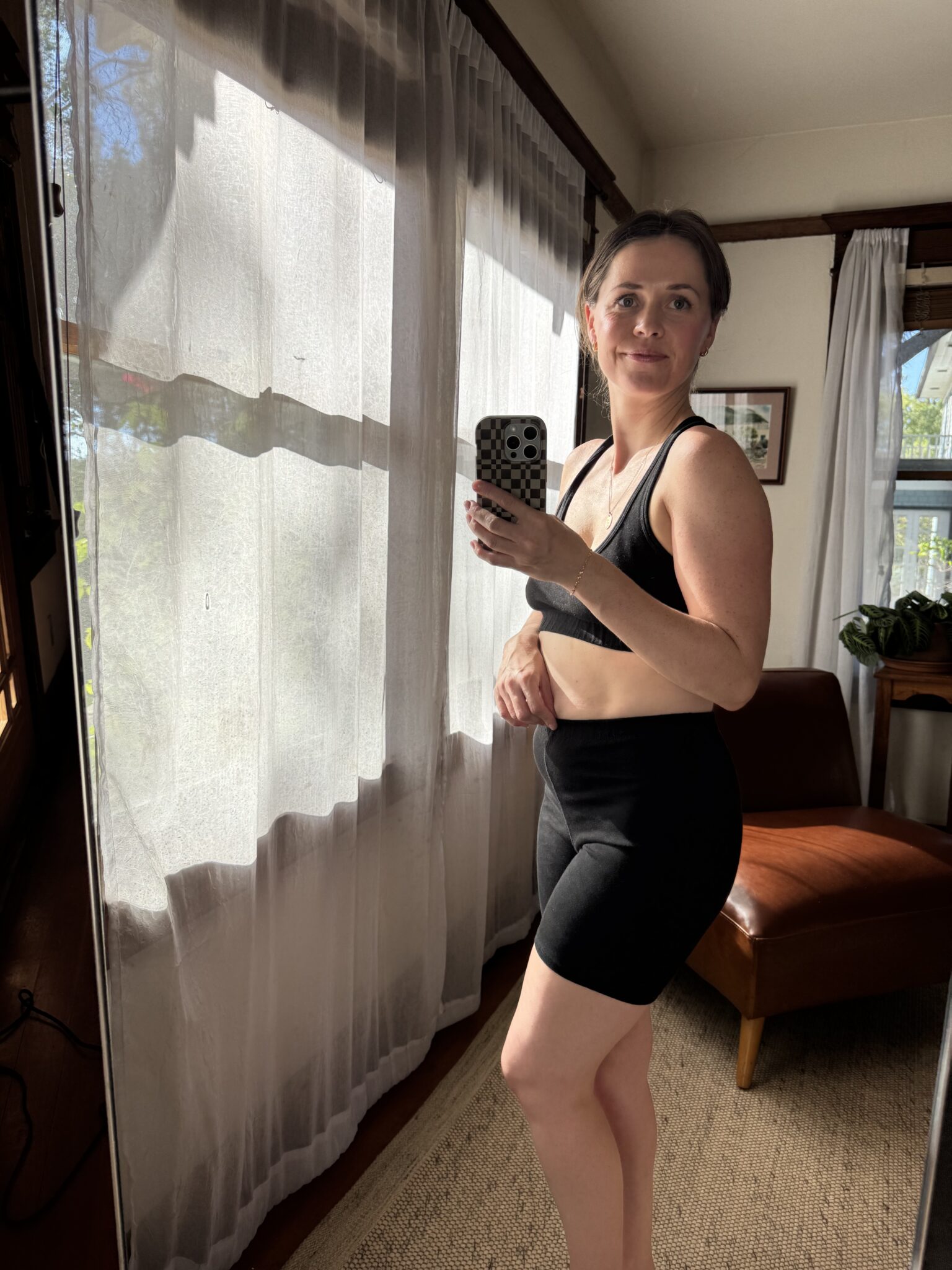 A woman in black workout clothes takes a mirror selfie in a sunlit room with sheer curtains and a brown chair.