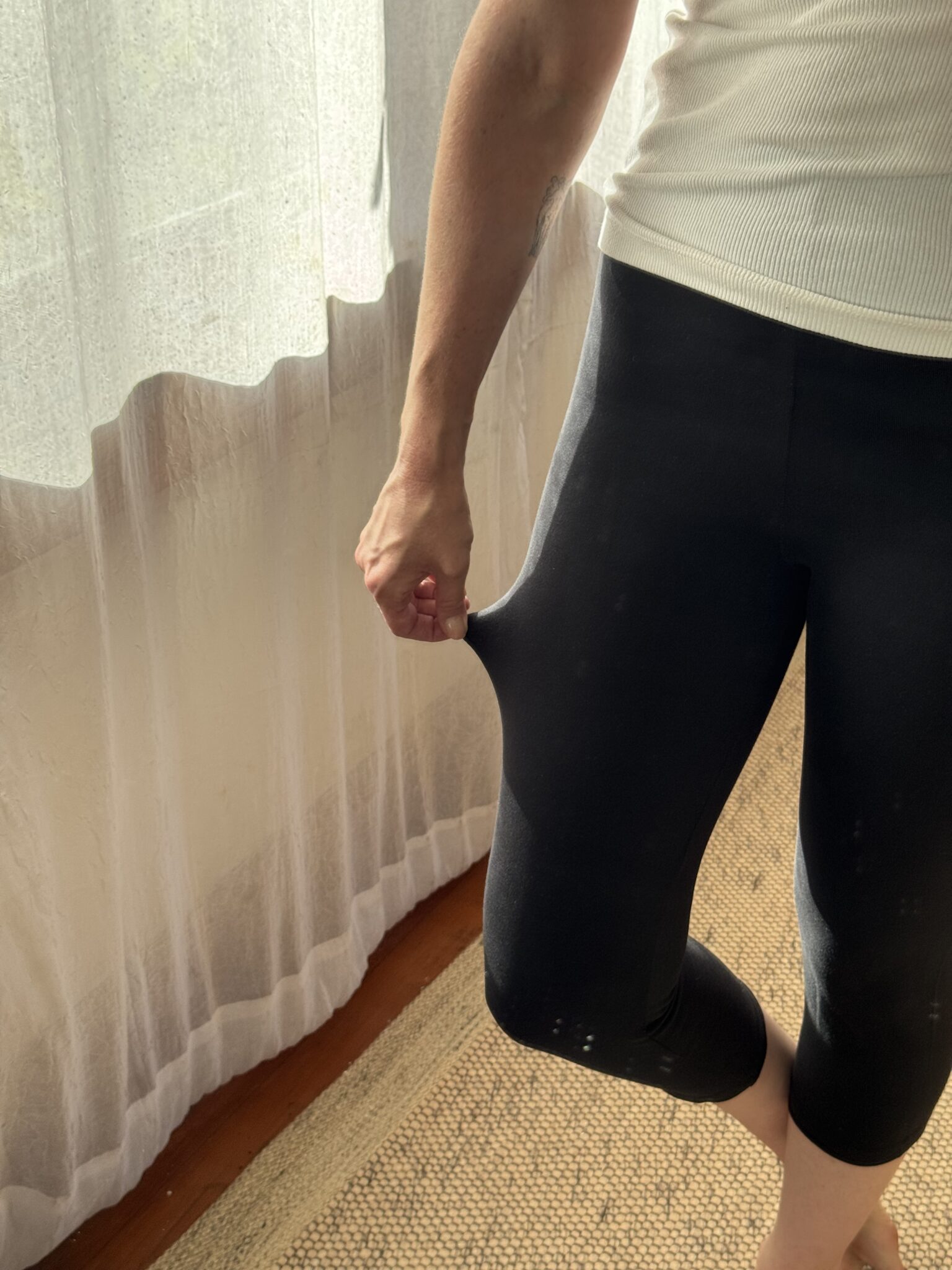 A person in a white top and dark leggings stands indoors, pulling the fabric of their leggings away from their thigh with one hand.