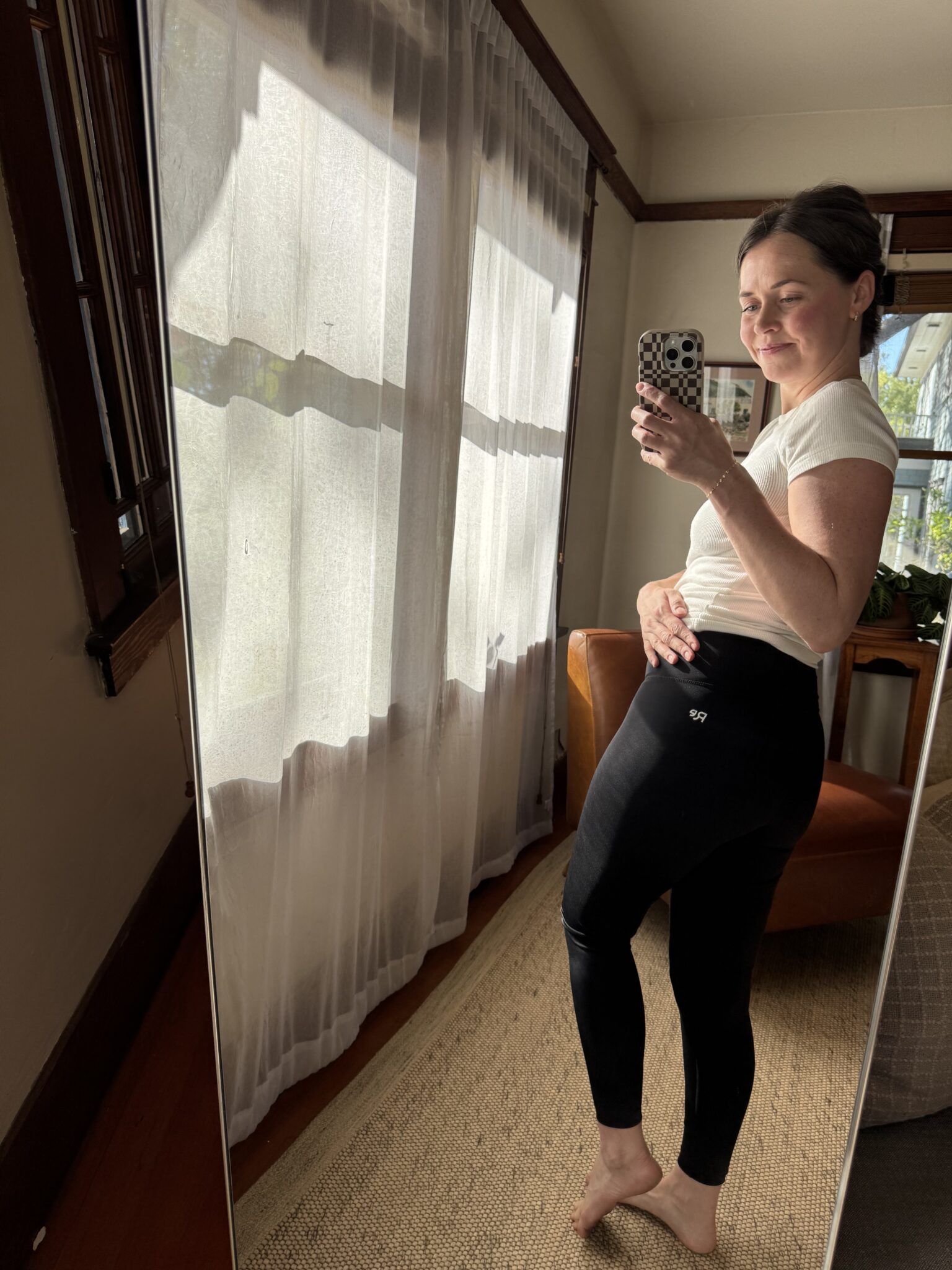 A person in a white t-shirt and black leggings stands barefoot in front of a mirror, taking a selfie in a sunlit room with curtains and a brown chair.