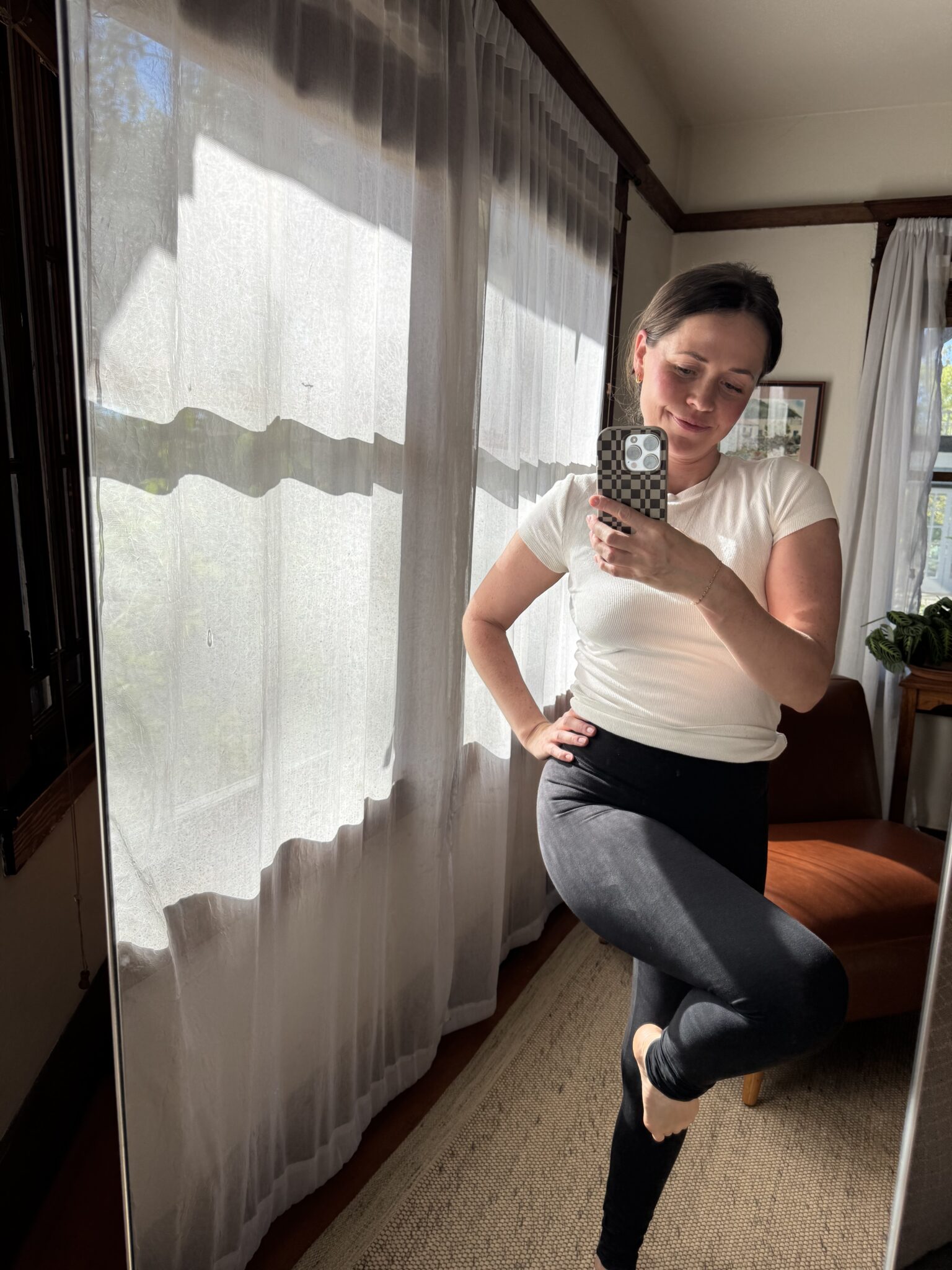 A person in a white shirt and black leggings takes a mirror selfie in a sunlit room with sheer curtains, a brown chair, and a plant visible in the background.