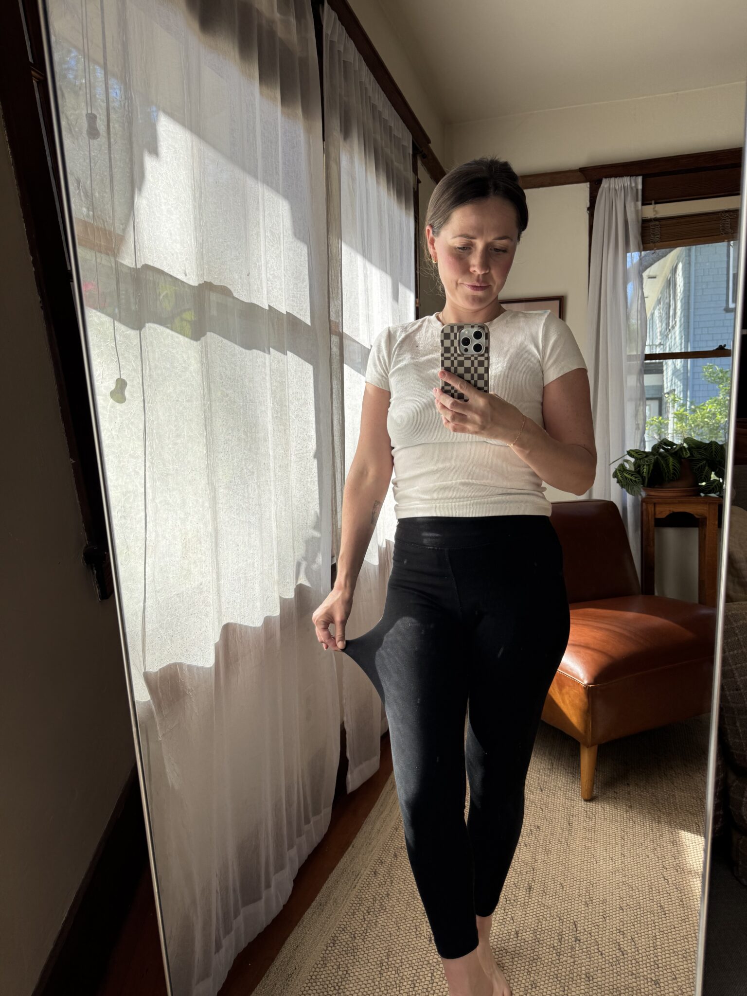 A person stands barefoot indoors, taking a mirror selfie. They wear a white top and black leggings, with sunlight streaming through sheer curtains behind them.