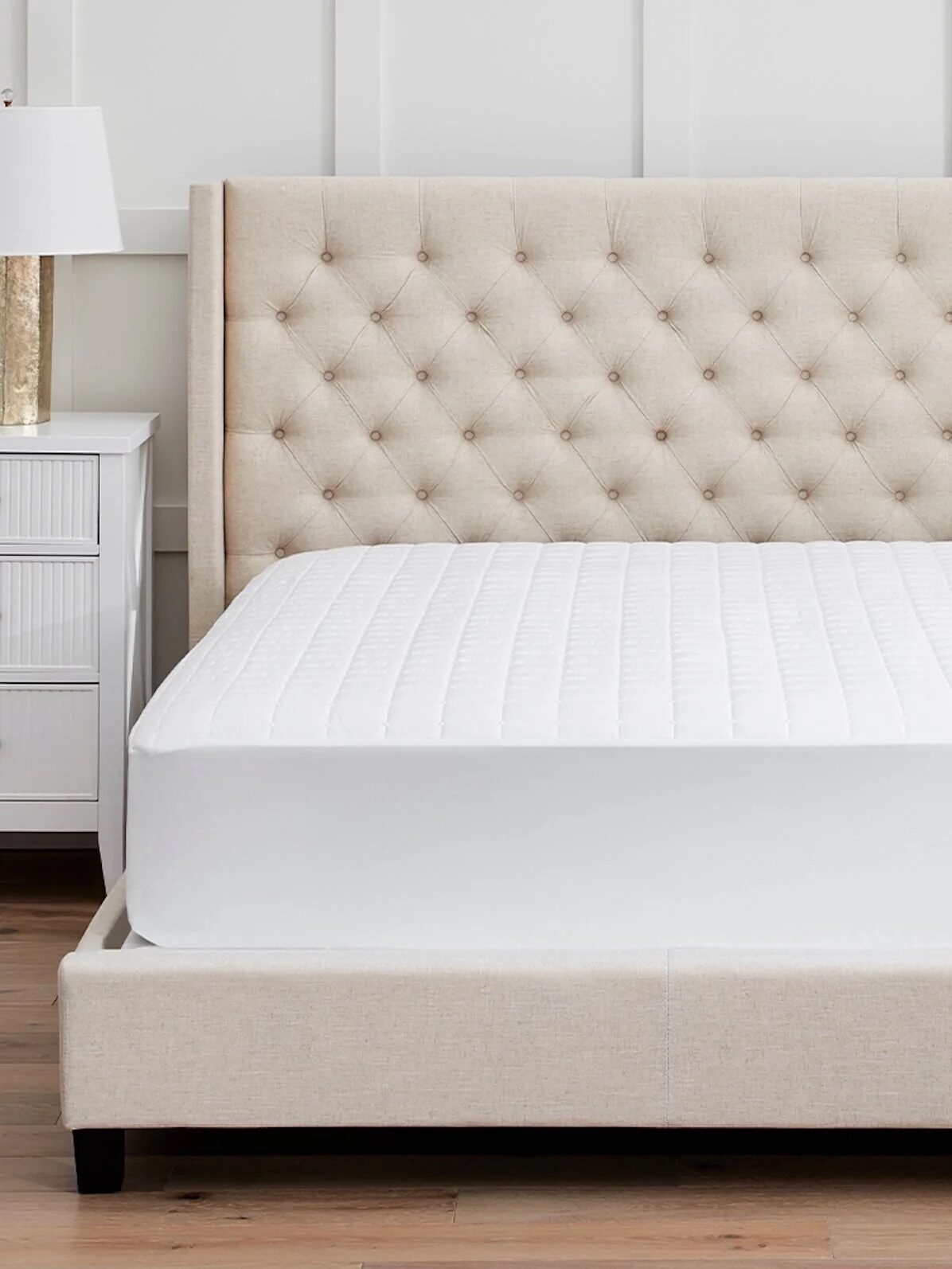 A neatly made bed with a beige tufted headboard, white mattress, and white bedding, flanked by two white nightstands with lamps.