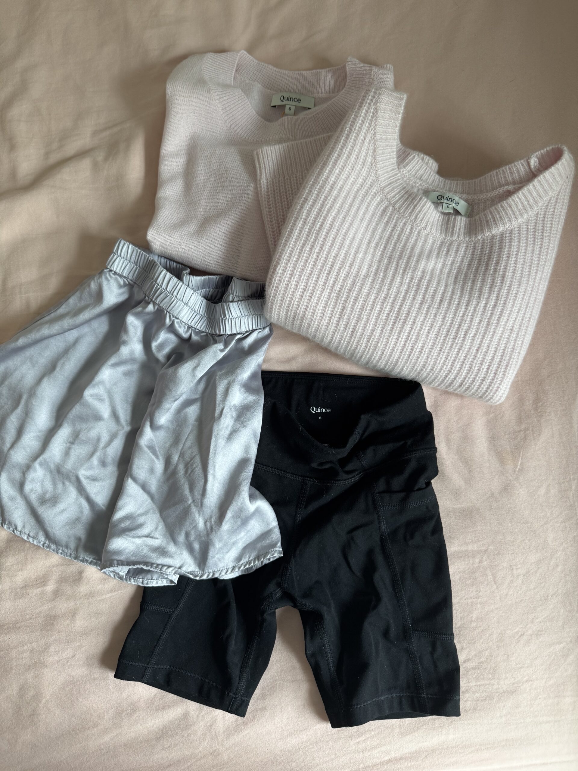 Two light pink sweaters, light blue athletic shorts, and black bike shorts are laid out on a light pink surface.