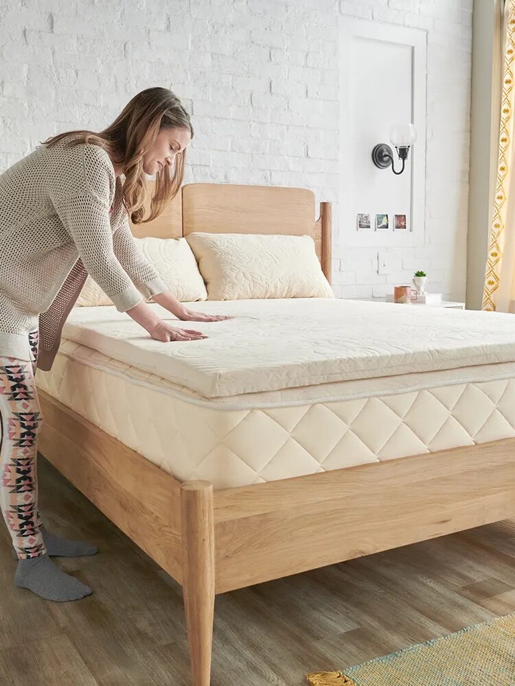 A woman arranges a quilt on a bed with a wooden frame in a bright bedroom featuring white brick walls, patterned curtains, and a small wall lamp.