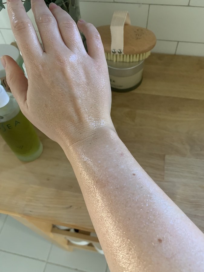 A hand and forearm with moisturized skin, resting on a wooden surface with a bottle and a container in the background.