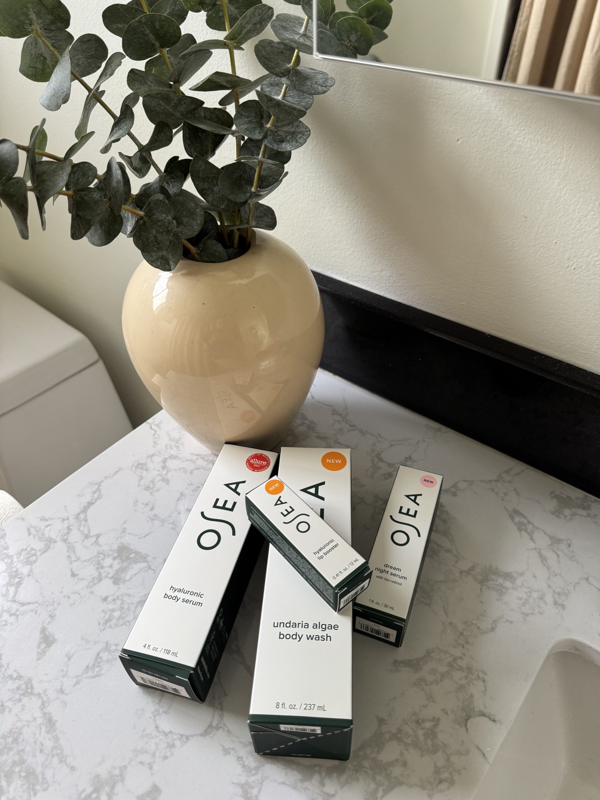 Three OSEA skincare product boxes are arranged on a marble countertop next to a beige vase with green foliage in a bathroom setting.