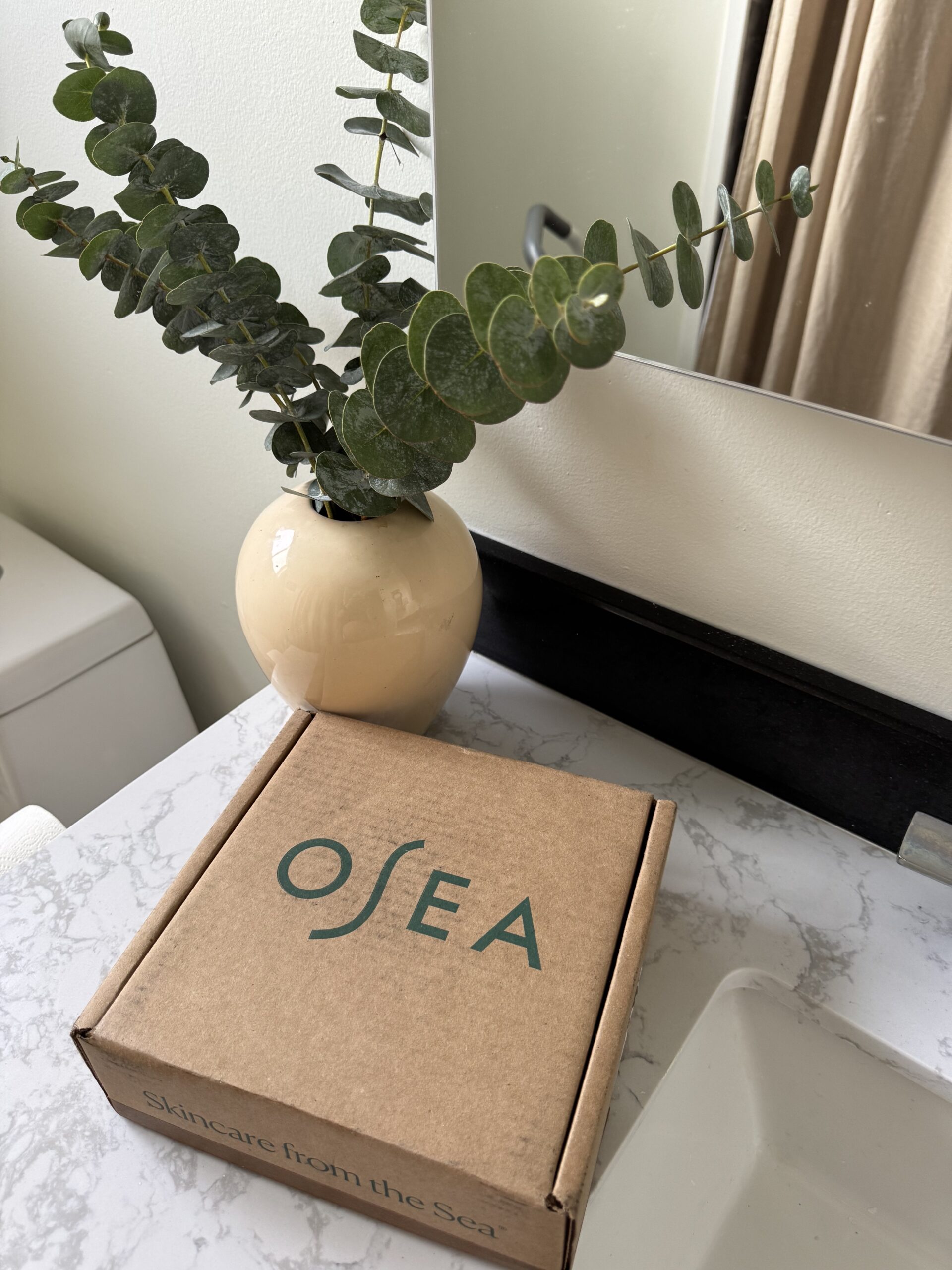 A cardboard Osea box labeled "Skincare from the Sea" sits on a marble countertop next to a beige vase with eucalyptus stems in a bathroom.