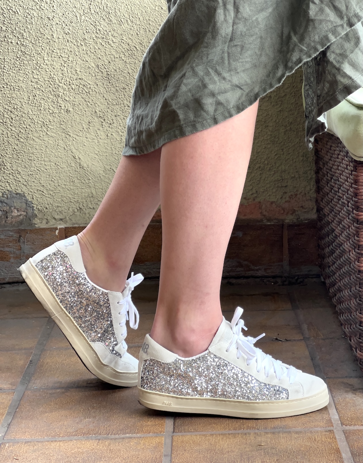Person wearing silver glitter sustainable sneakers and a green skirt, sitting with legs crossed on a tiled floor next to a wicker basket.