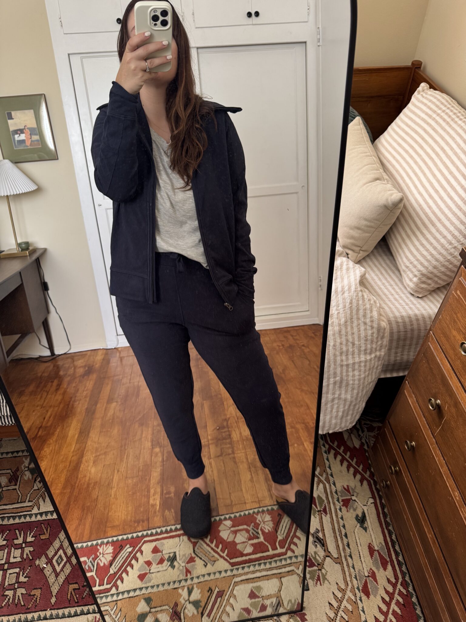 A person stands in front of a mirror taking a selfie, wearing a black jacket, gray shirt, black pants, and slippers in a bedroom with wooden floors and neutral decor.