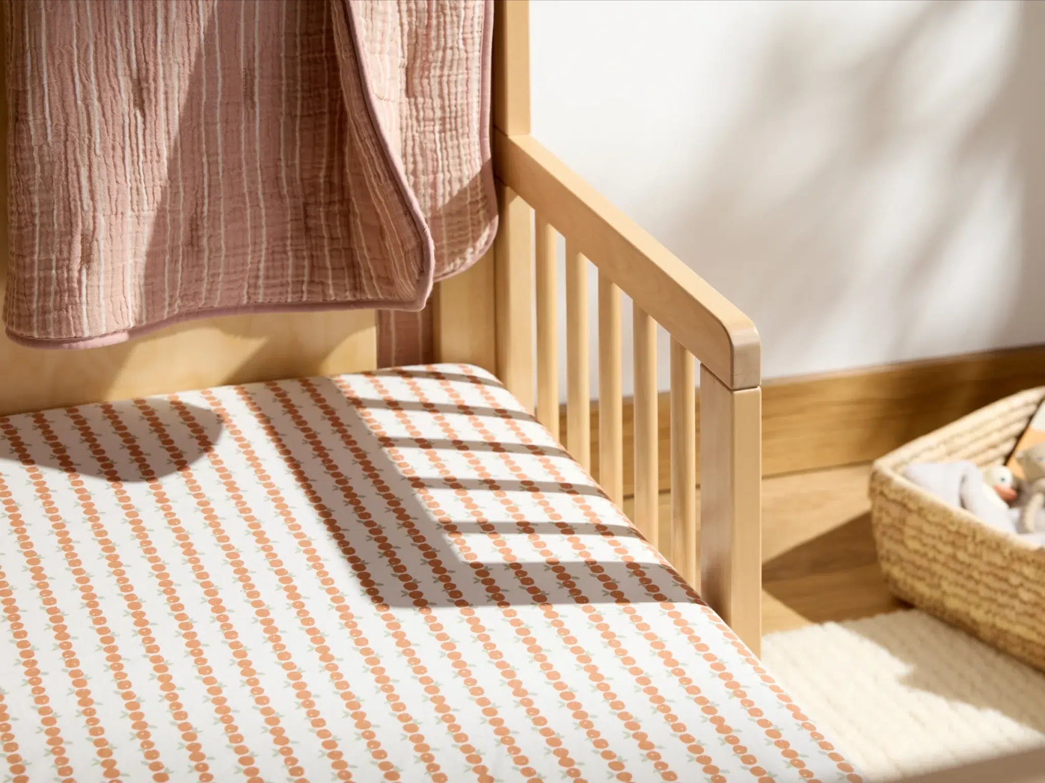 A wooden crib with a patterned sheet and a pink blanket draped over the side, next to a woven basket on the floor.