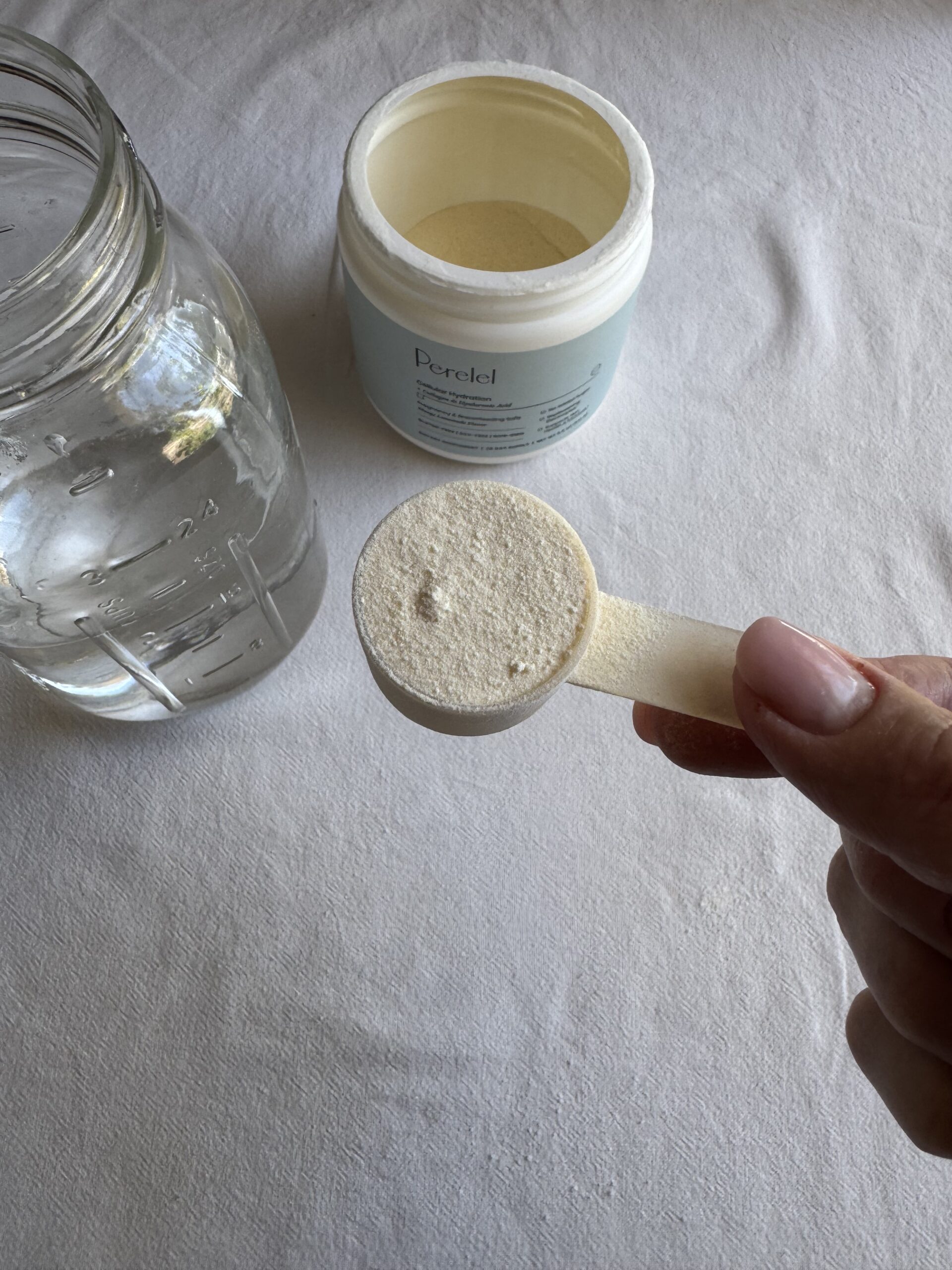 A hand holds a scoop of powdered supplement above a jar of water and an open container labeled "Perlel" on a white surface.
