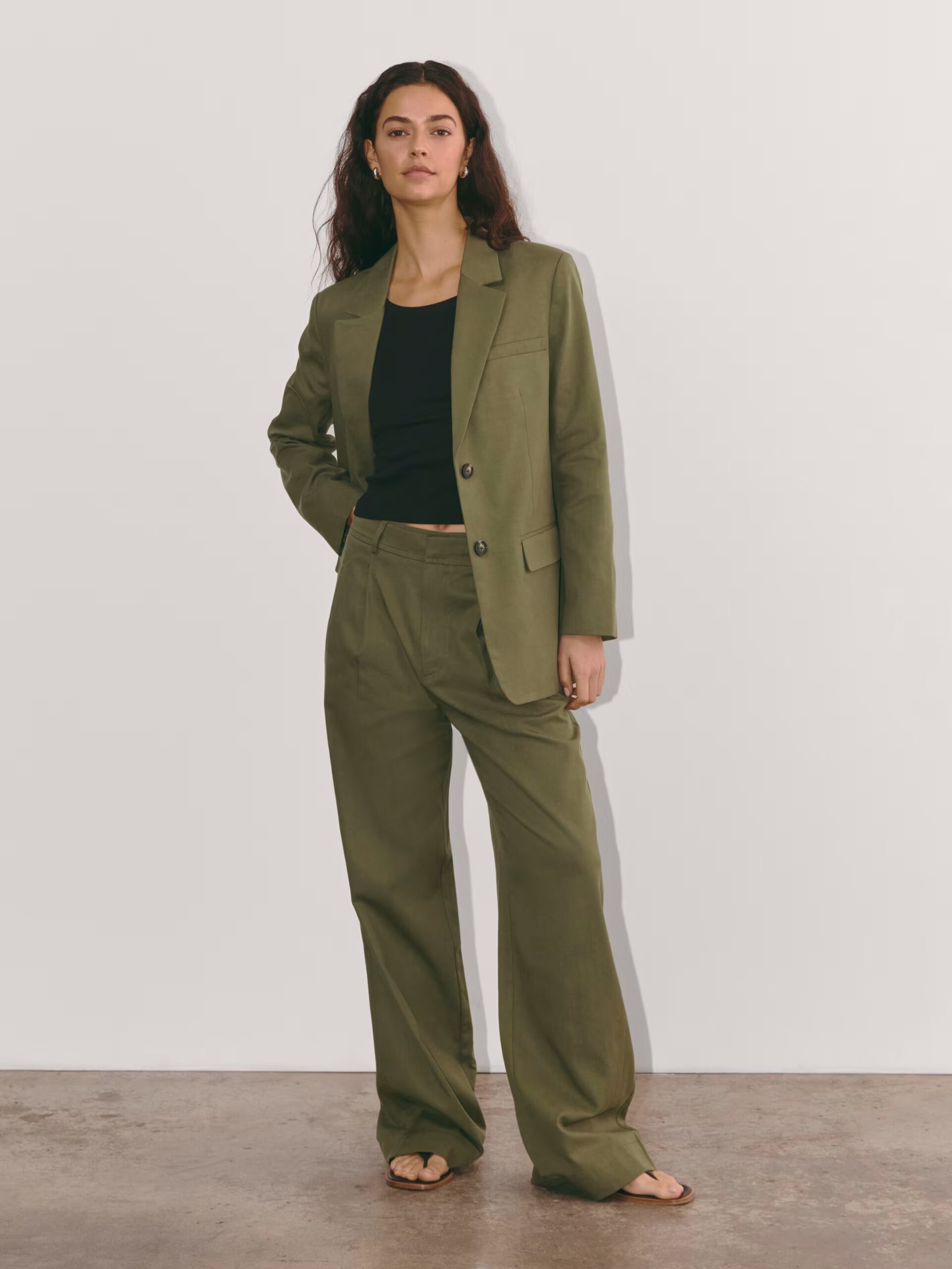 A person stands against a plain background wearing an olive green suit with a black top and sandals.