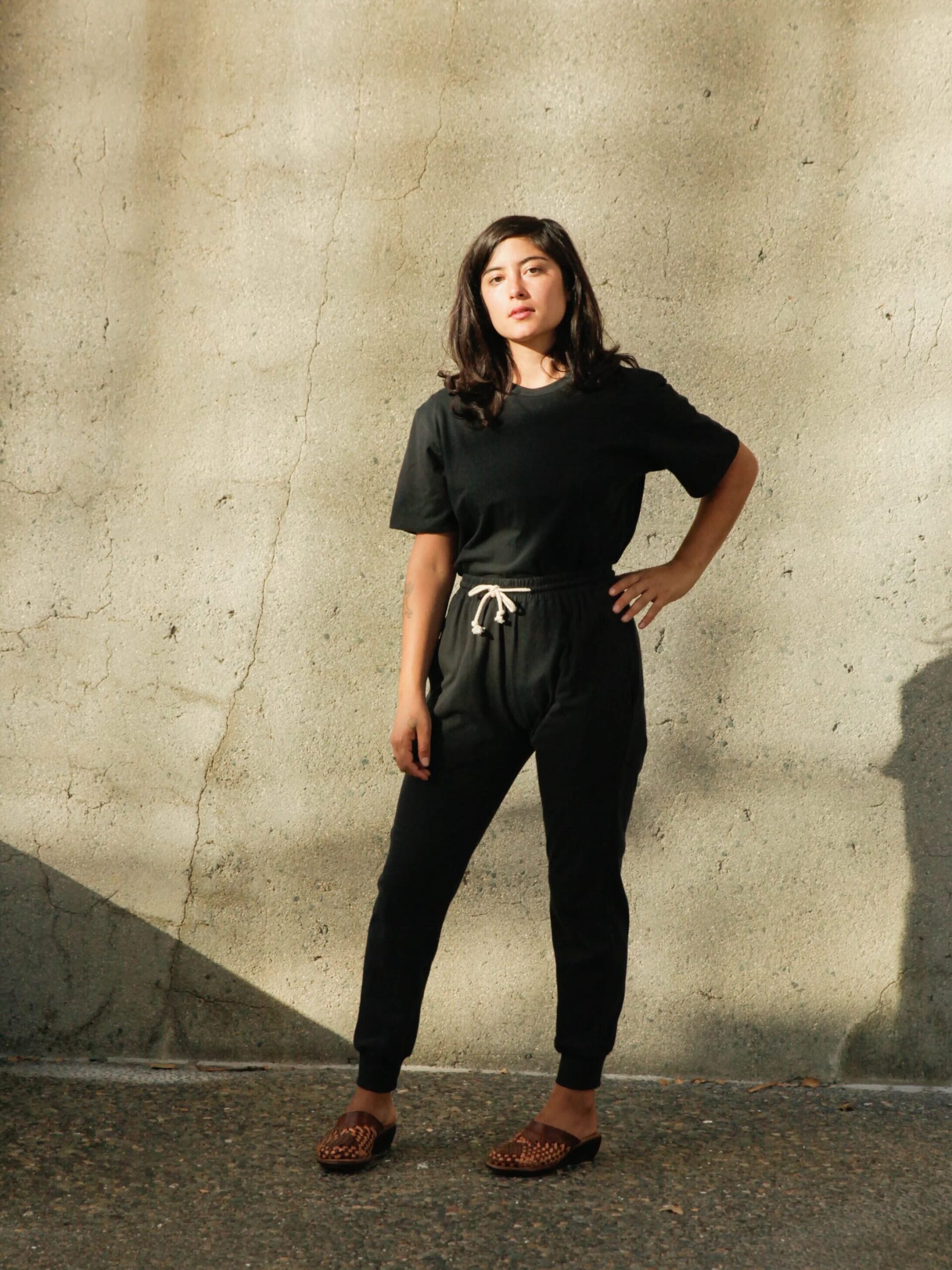 Person in black outfit stands against a sunlit textured wall, with one hand on hip. Wearing slippers, the individual looks towards the camera.