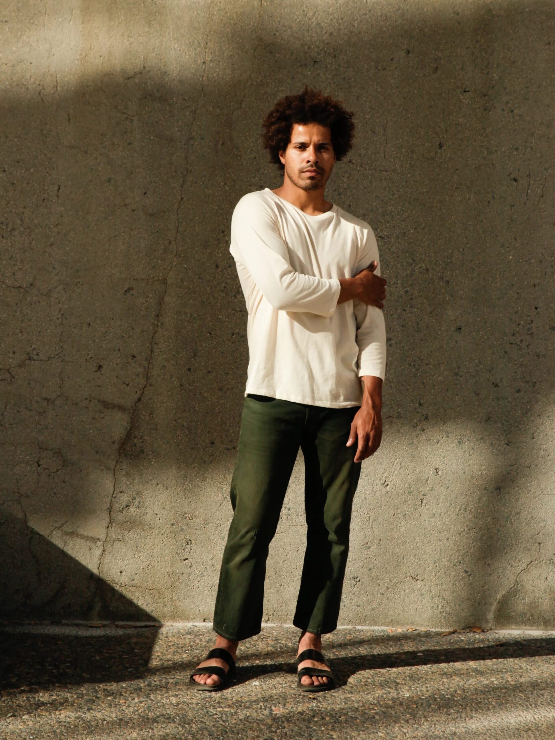 A person with curly hair stands against a textured wall, wearing a long-sleeve white shirt, green pants, and sandals, with one arm crossed over their chest.