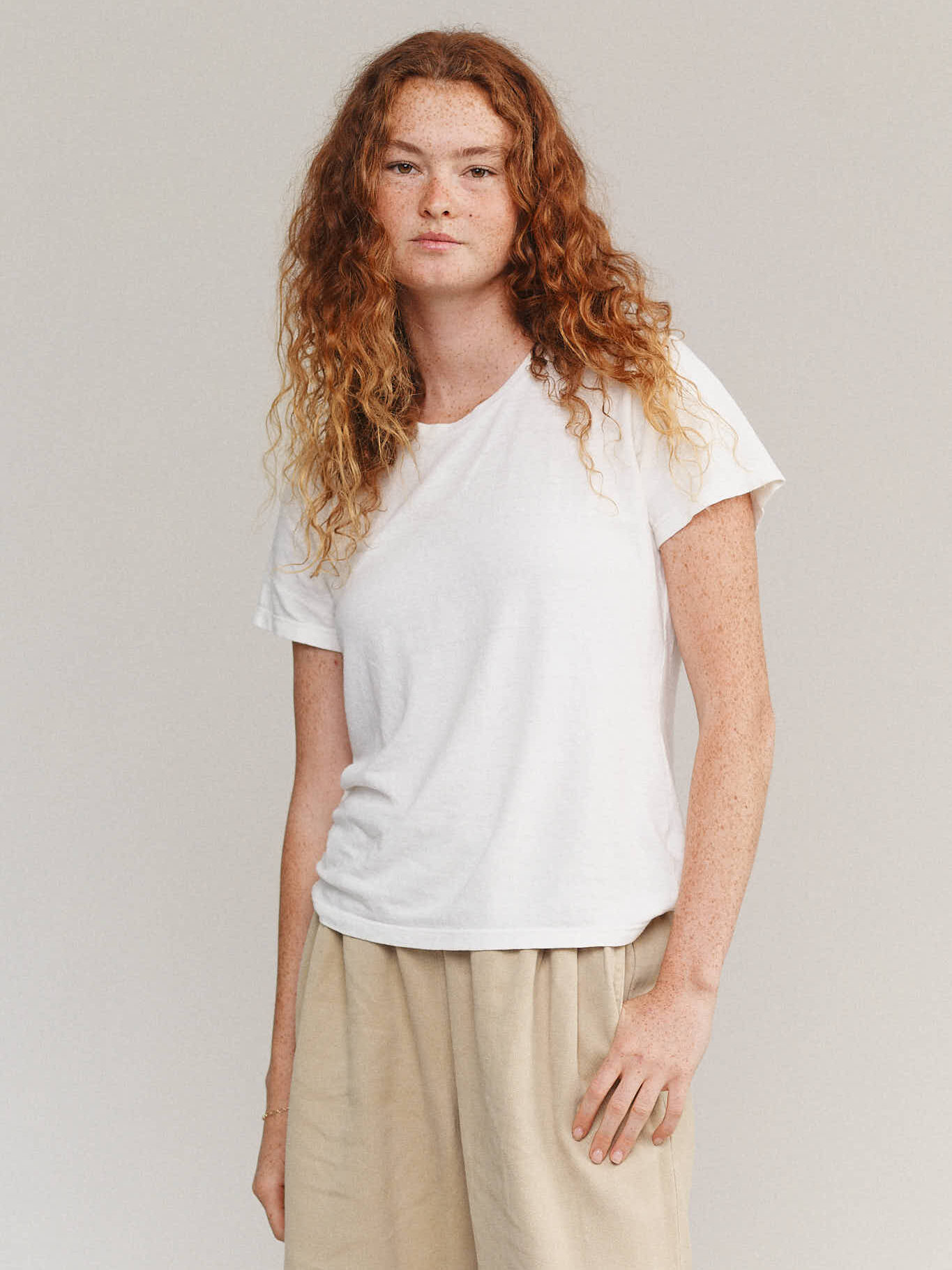 Person with long curly hair wearing a white t-shirt and beige pants stands against a plain background.