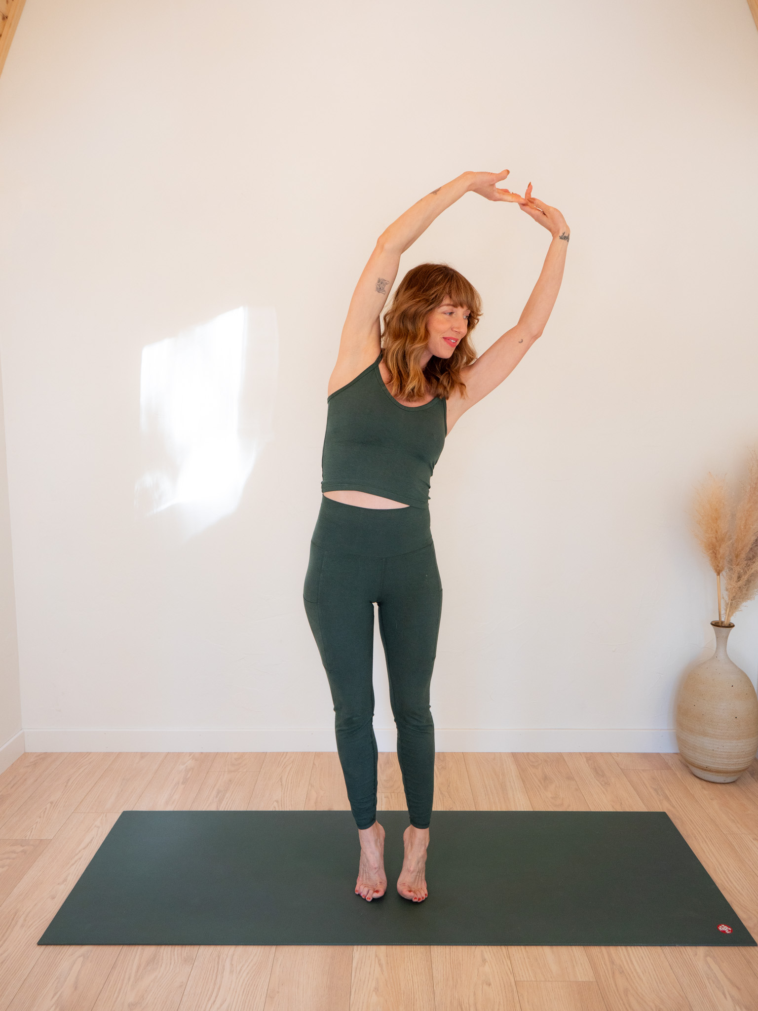 A woman in a dark green workout outfit performs a standing side stretch on a yoga mat in a bright room.