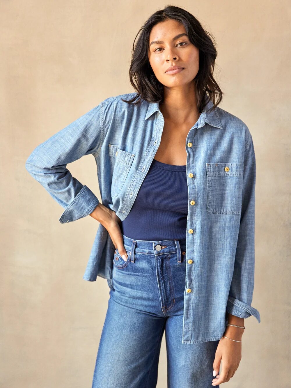 Person wearing a blue denim shirt over a dark top and jeans, standing against a neutral background.