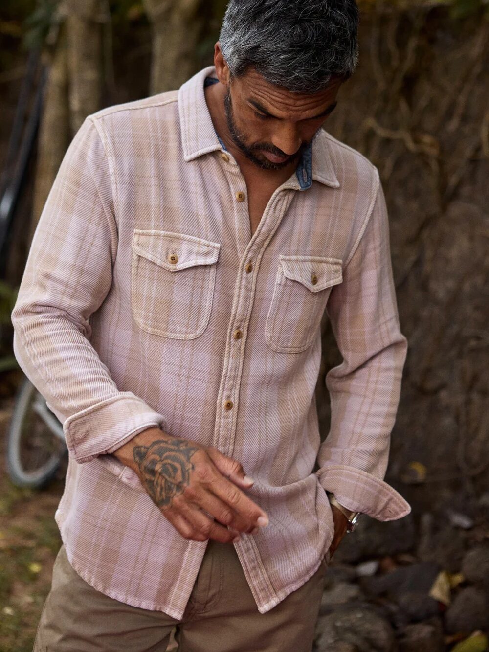 Man with gray hair stands outdoors, wearing a light plaid shirt and brown pants. He has a tattoo on his left hand and appears to be looking down. A bicycle is partially visible behind him.